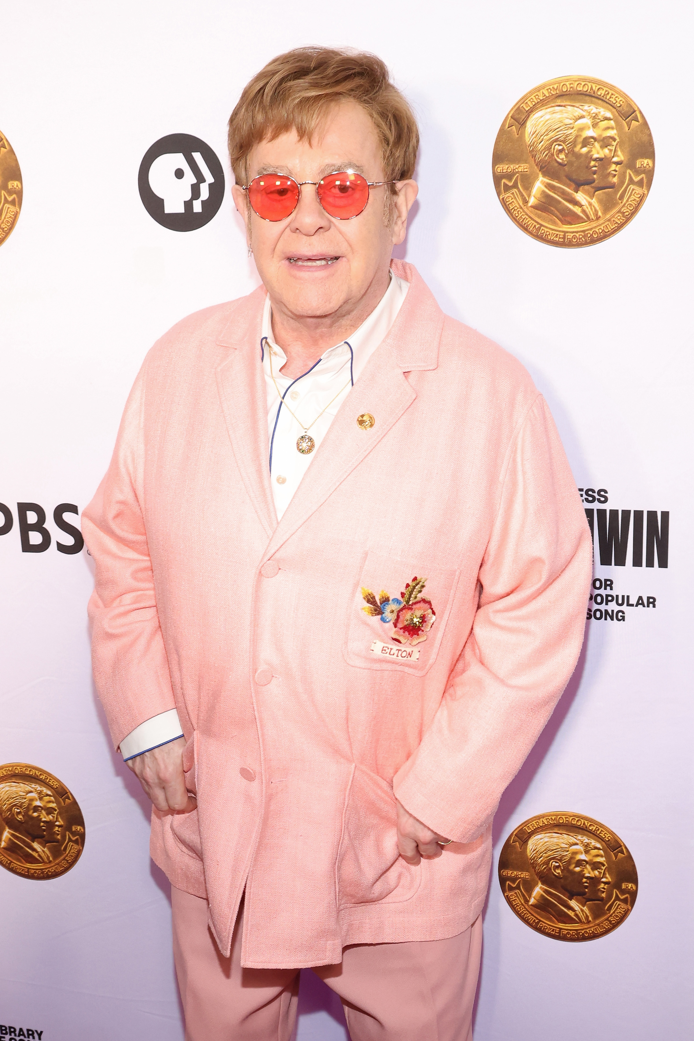 Elton John poses on the red carpet, wearing a light-toned blazer with a floral badge, glasses with tinted lenses, and a medallion necklace