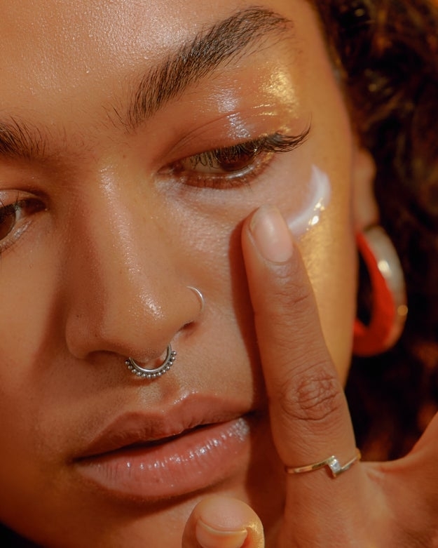 Close-up of a person applying facial cream near their eye with a finger. They have a septum piercing, hoop earrings, and curly hair