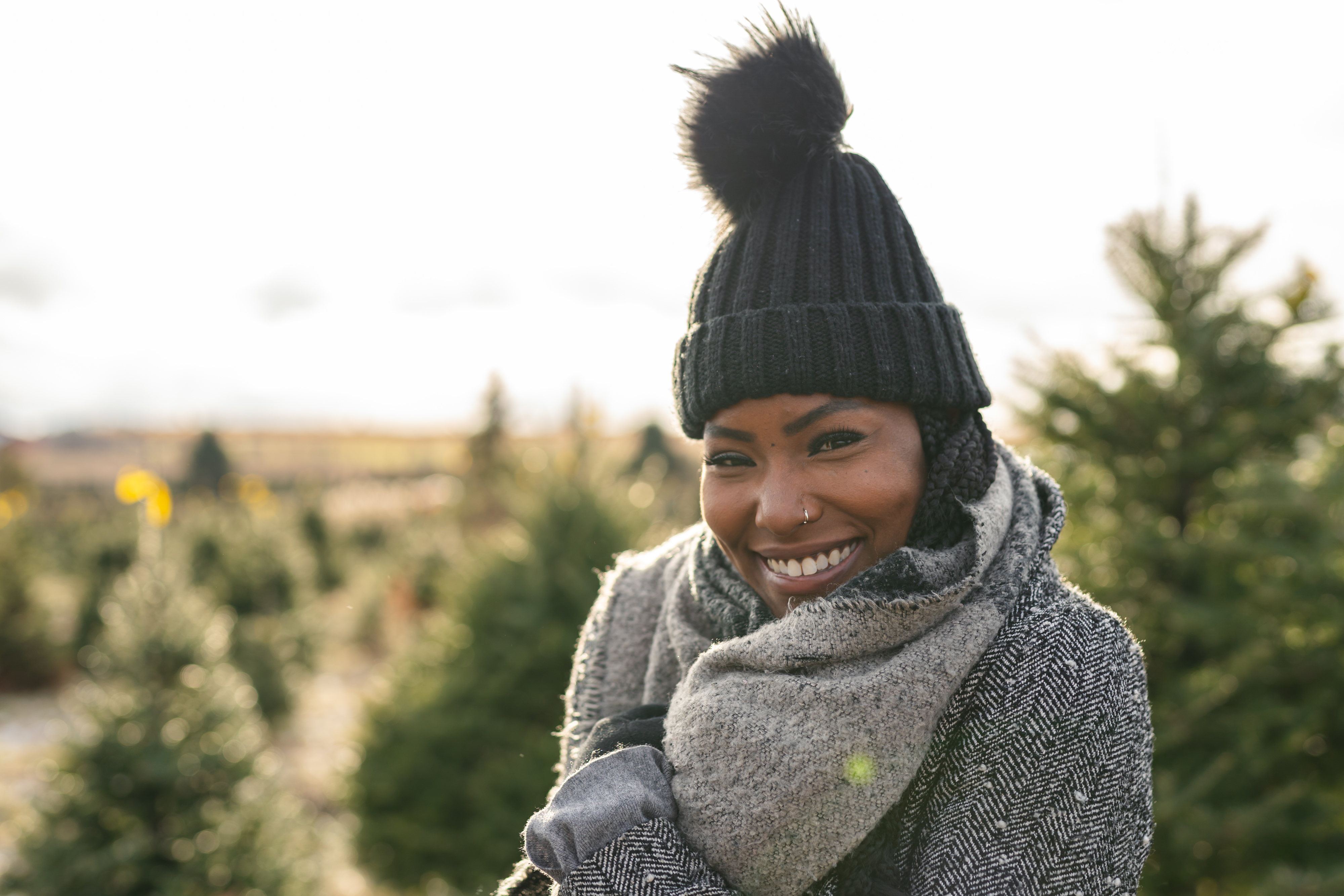 A person wearing a knit hat with a pom-pom and a cozy scarf smiles warmly while standing amidst Christmas trees at an outdoor tree lot