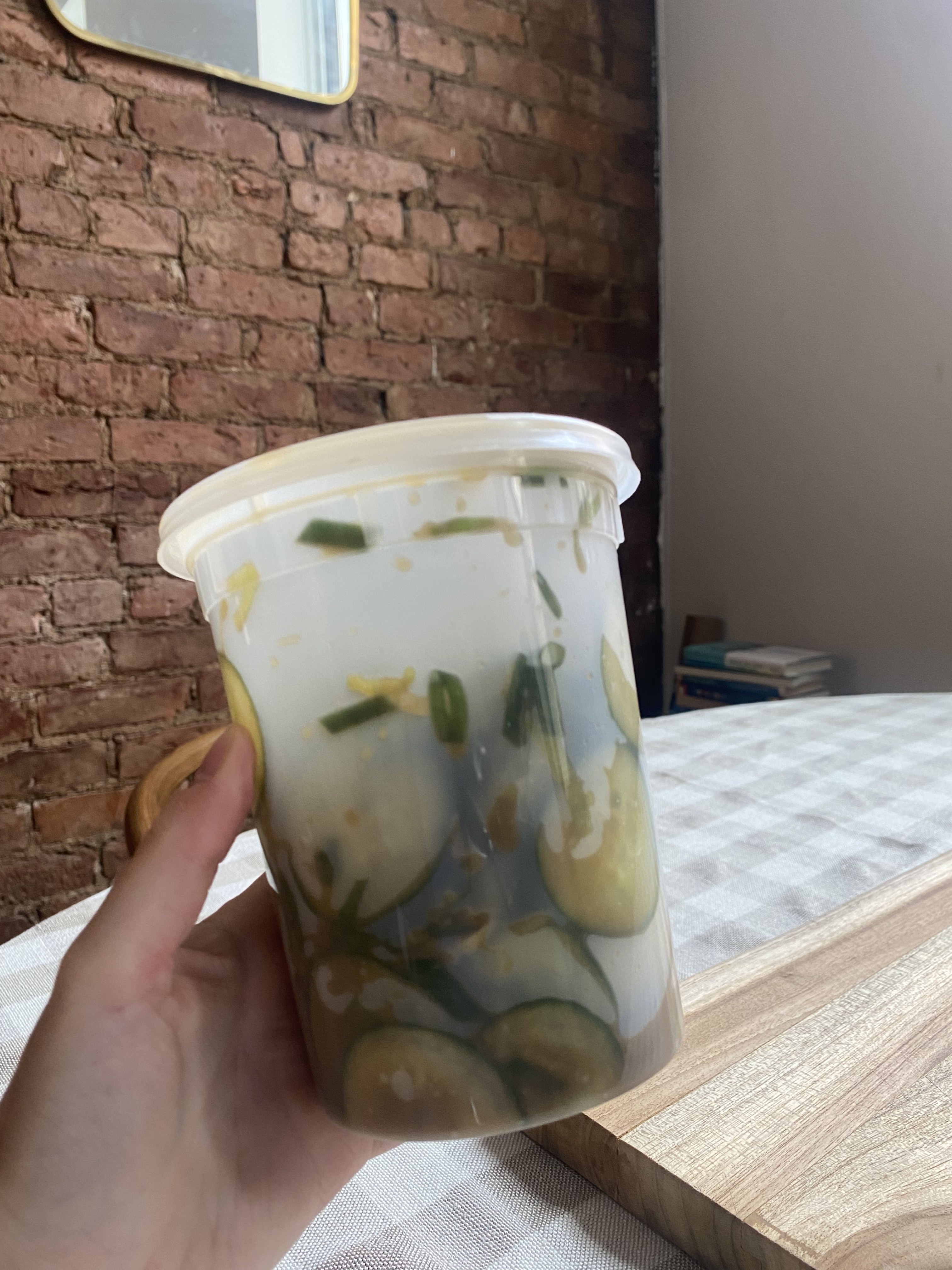 A hand holds a plastic container filled with sliced cucumbers, herbs, and liquid against a rustic brick wall