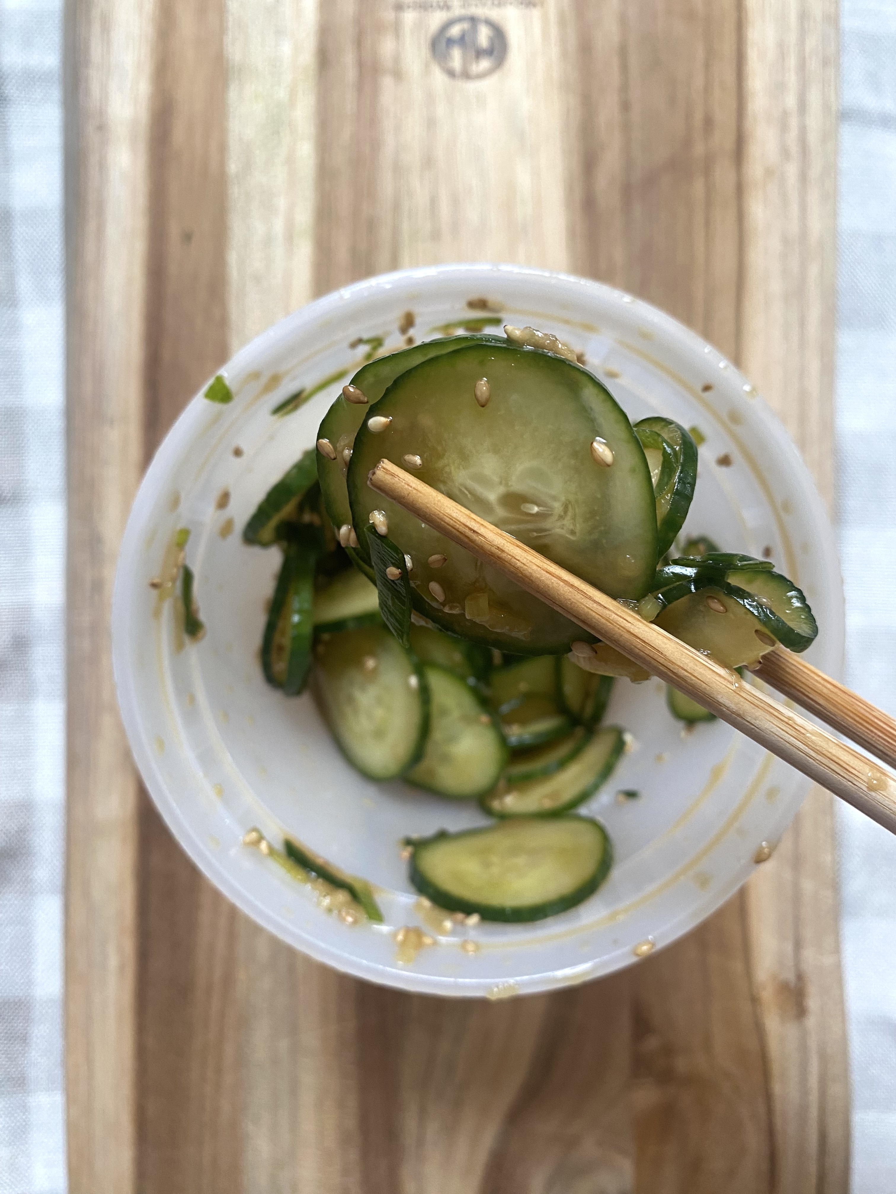 Chopsticks holding a sesame-covered cucumber slice above a container of cucumber salad. The bowl rests on a wooden surface