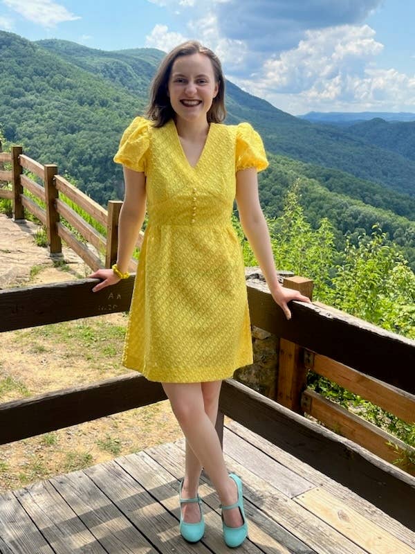 The author smiling in a patterned dress standing on a wooden deck with mountains and lush green trees in the background