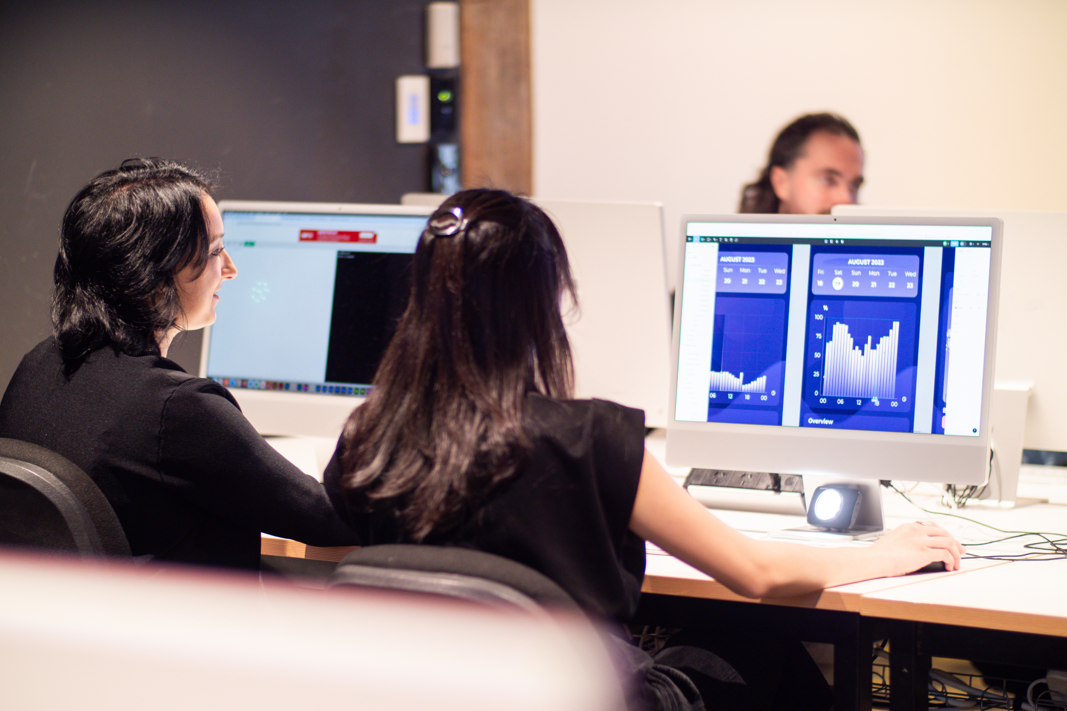 Two people are working on desktop computers displaying graphs and charts in an office environment. Another person is seen in the background