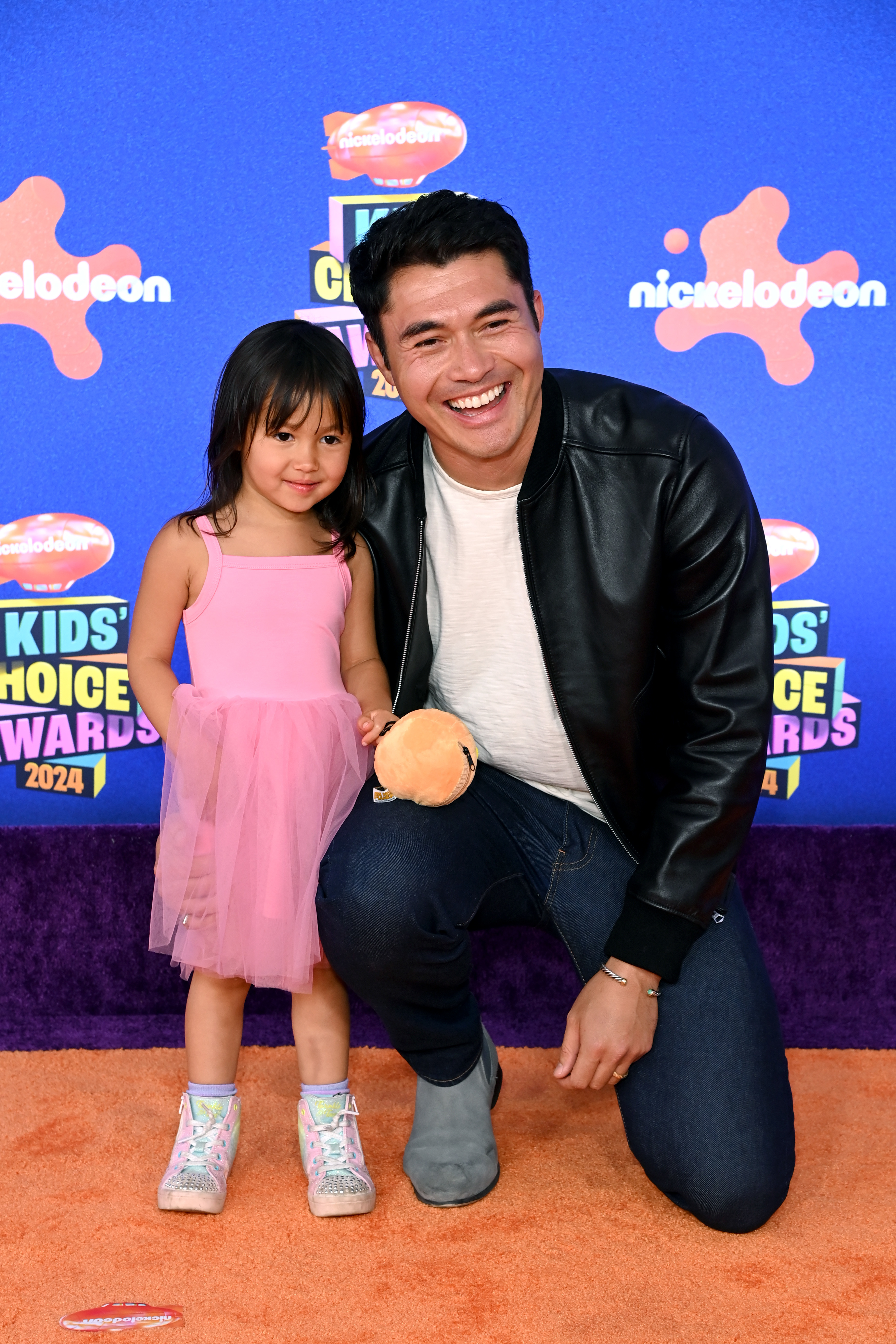 Henry Golding, in a casual outfit with a black jacket and jeans, kneels next to his daughter, who wears a pink dress, on the orange carpet at the Kids' Choice Awards 2024