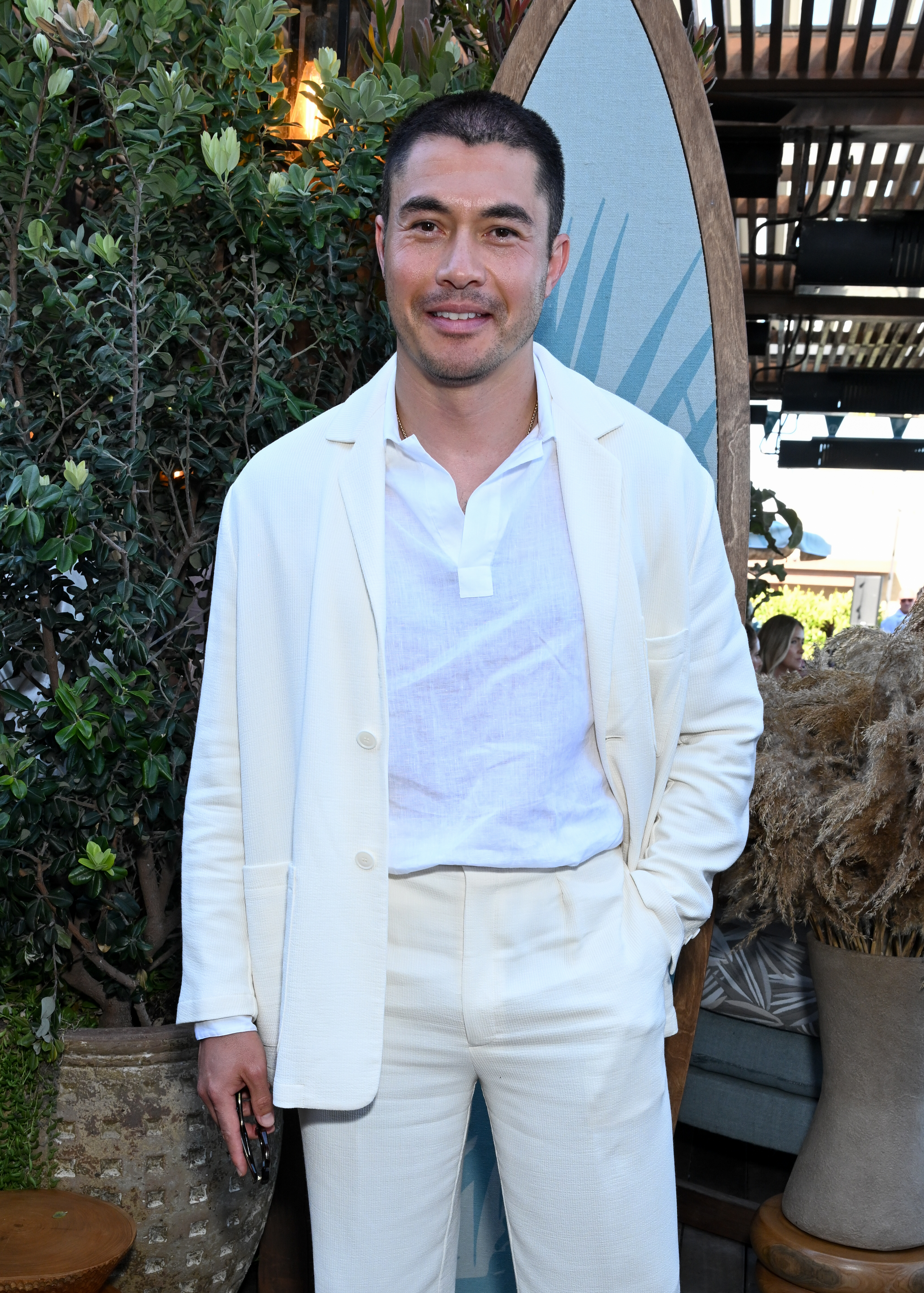 Henry Golding in a light suit and open-collar shirt, standing outdoors with greenery in the background