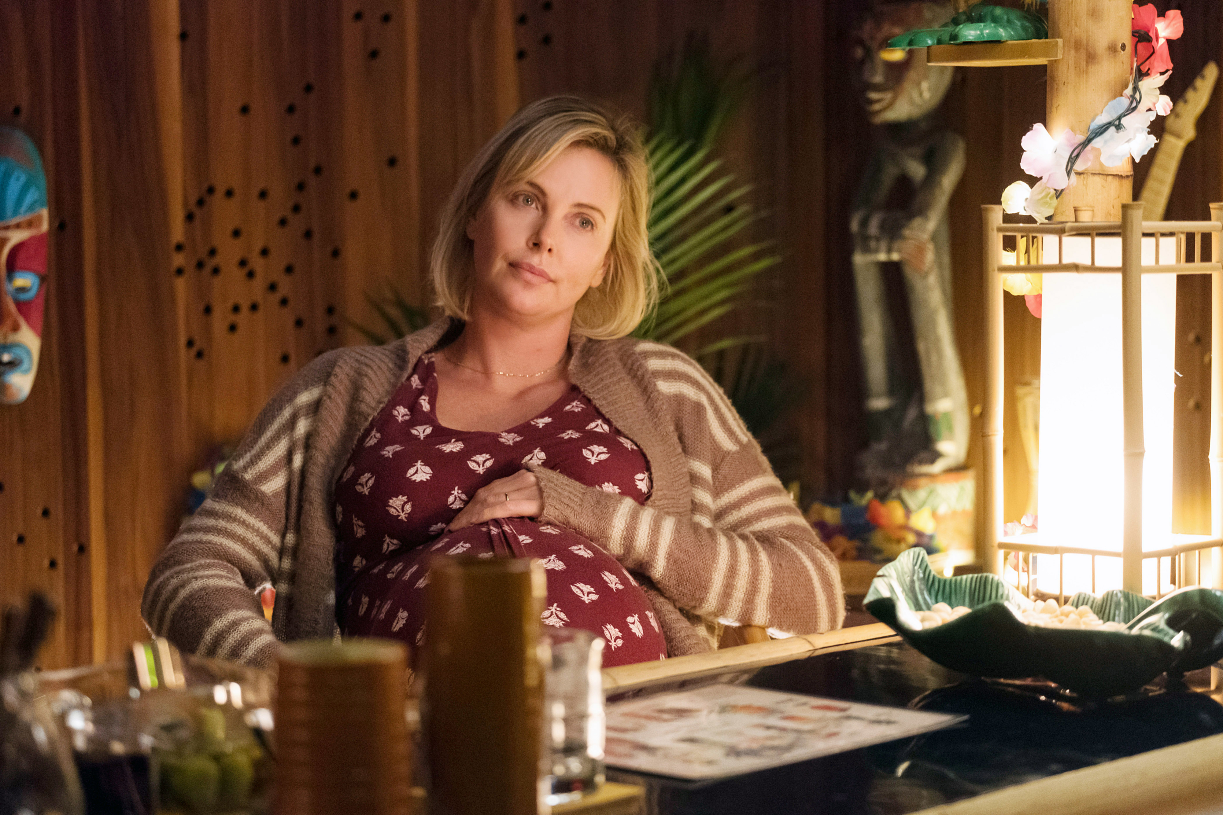 Charlize Theron, visibly pregnant, wears a patterned dress and striped cardigan while sitting at a table in a cozy, warmly lit room in "Tully"