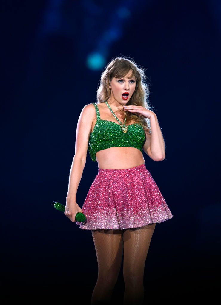 Taylor Swift performs on stage wearing a sparkly crop top and a glittery skirt, holding a microphone