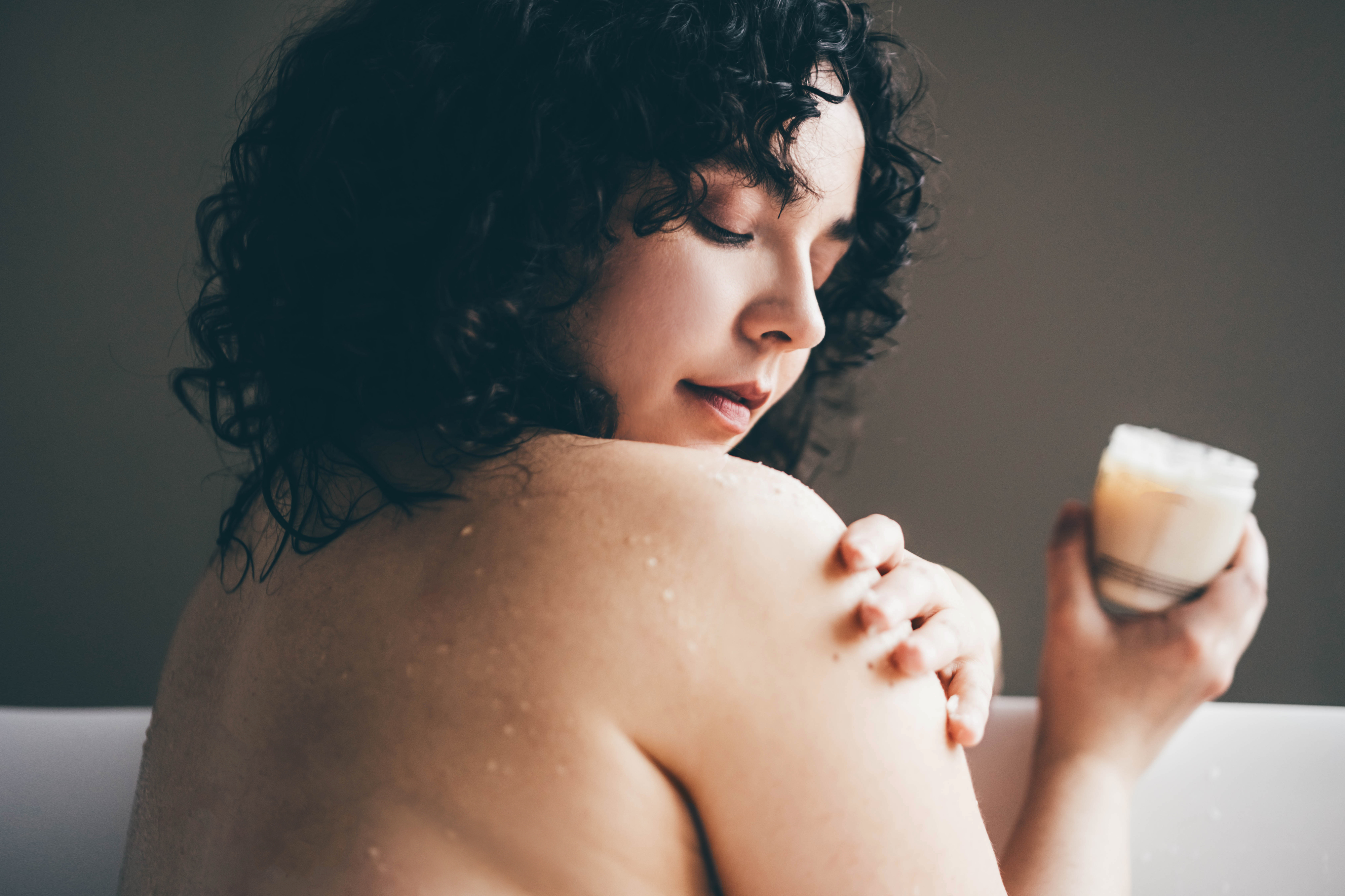 A woman with curly hair applies cream to her shoulder while holding a jar of lotion, showcasing a beauty and skincare routine