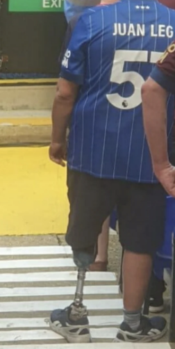 A person with a prosthetic leg stands wearing a striped shirt that says "Juan Leg 5" on the back. They are near a crosswalk