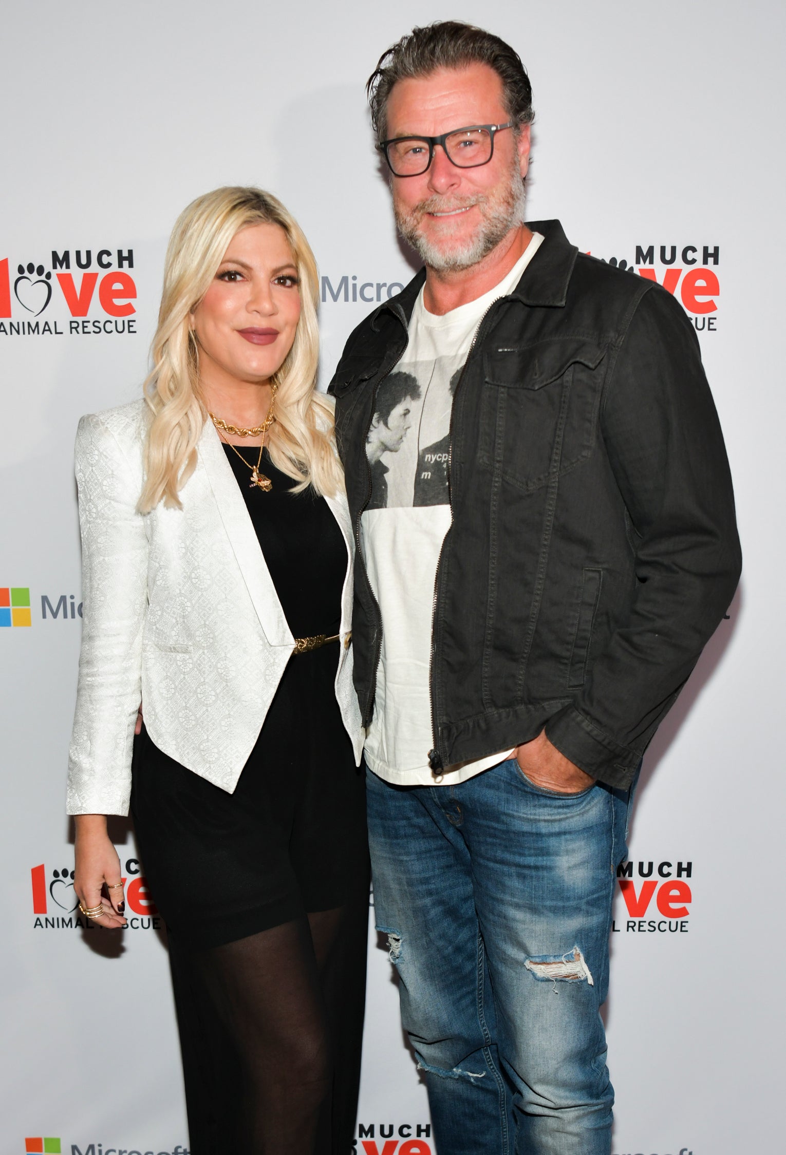 Tori Spelling in a white blazer and black dress, standing with Dean McDermott in a casual jacket and jeans at a Much Love Animal Rescue event