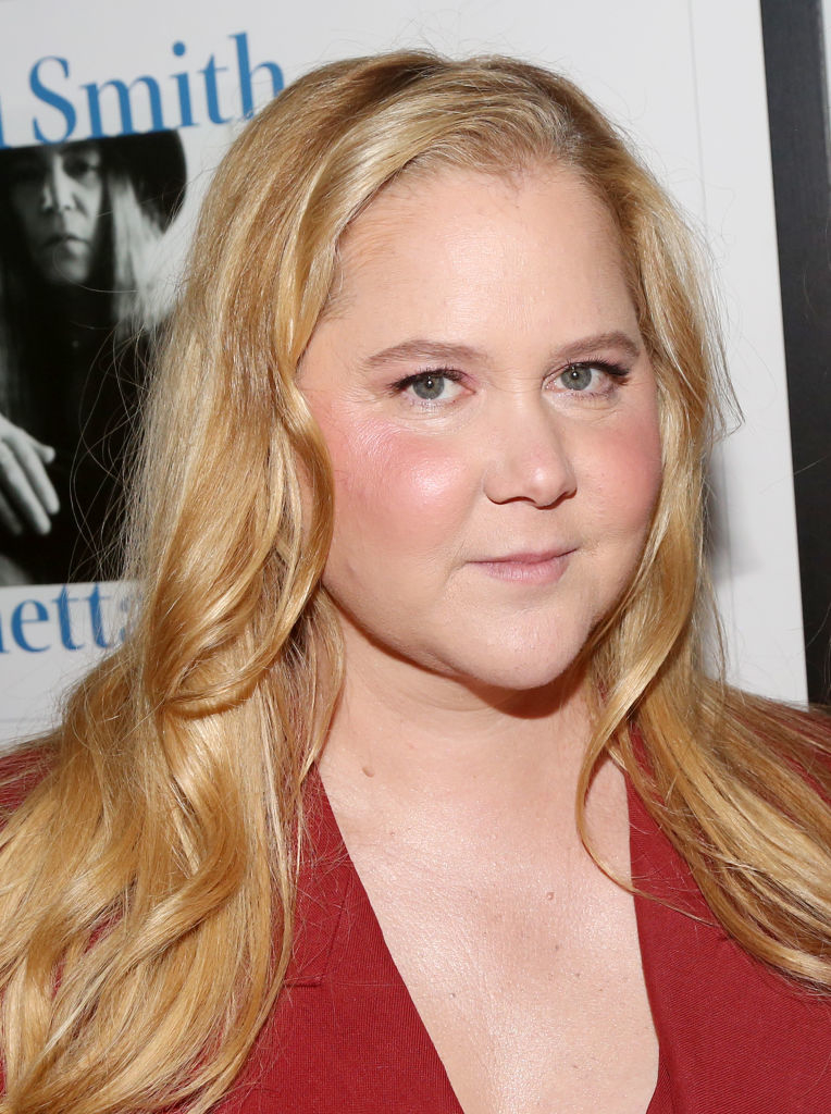 Amy Schumer at an event, wearing a smart outfit, with a poster in the background