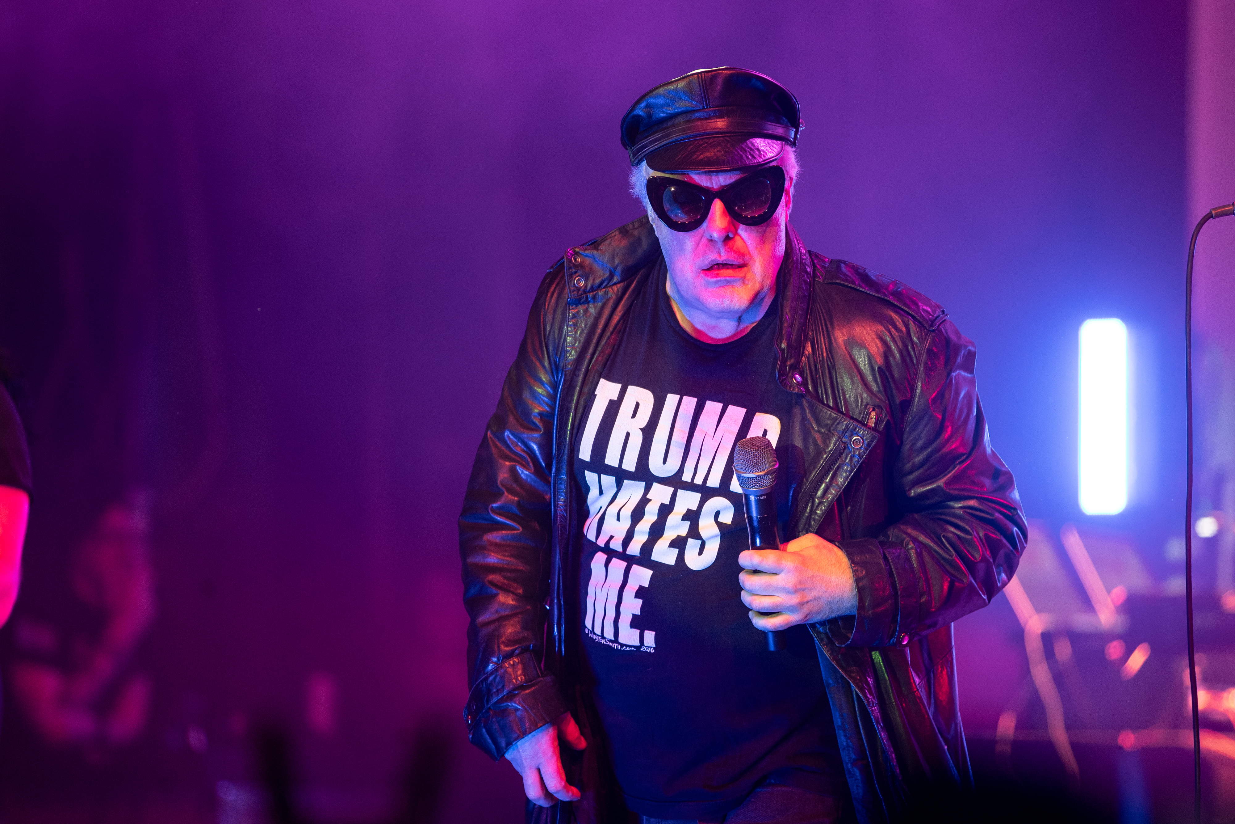 Jello Biafra wearing sunglasses, black leather cap and jacket, and a t-shirt with bold text &quot;TRUMP HATES ME.&quot; He is holding a microphone on stage