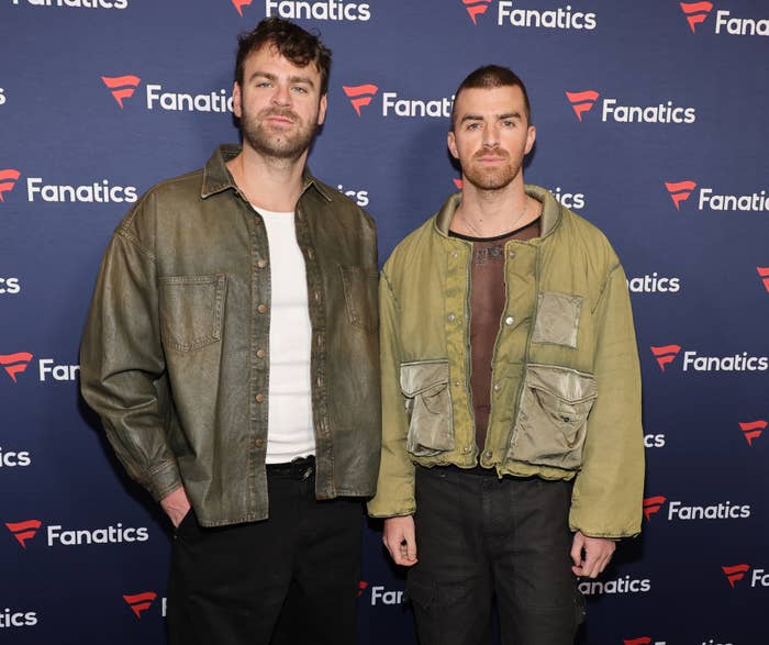 Drew Taggart and Alex Pall stand in front of a Fanatics backdrop at a celebrity event. Both are wearing casual, rugged jackets