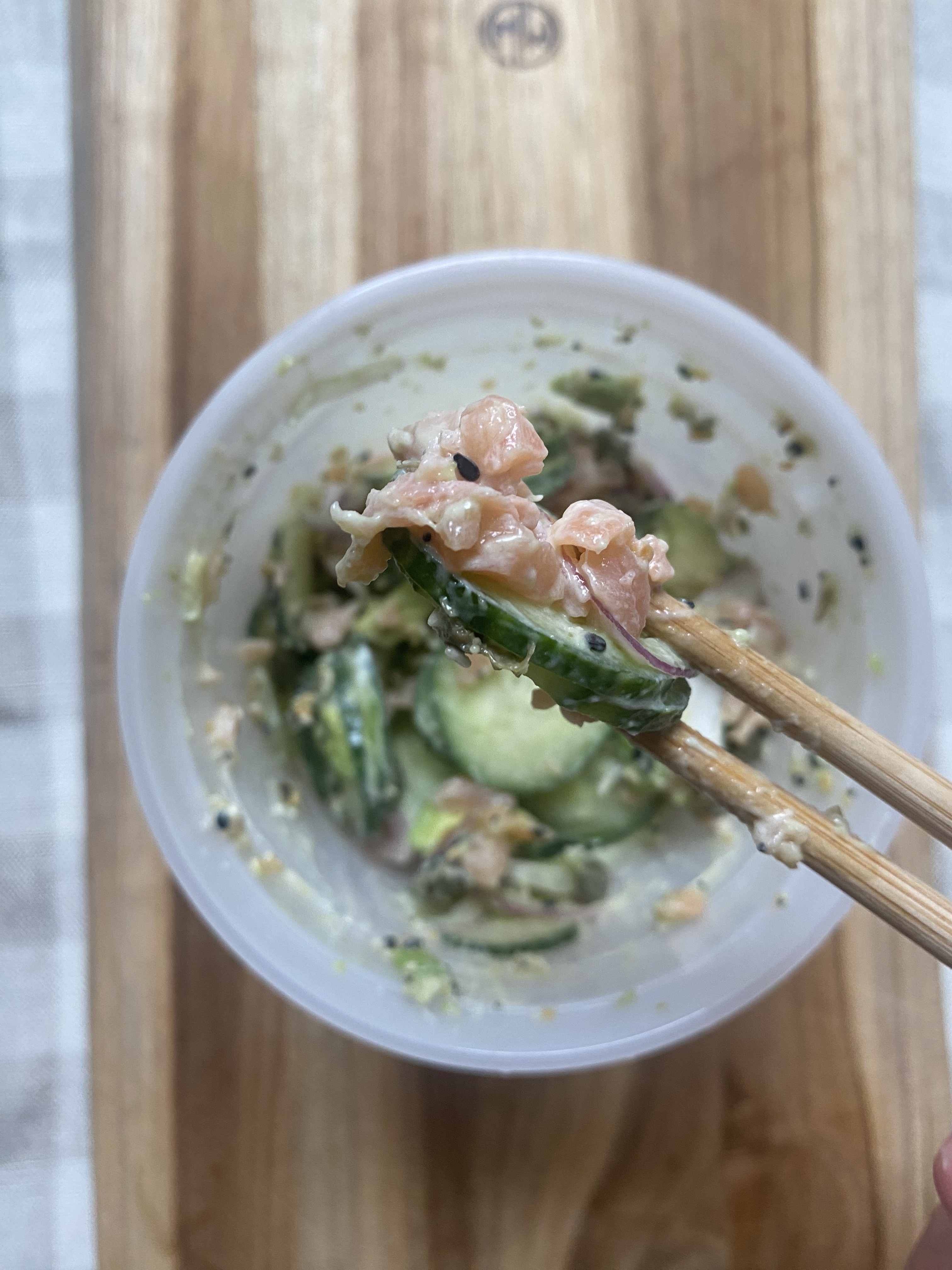 Bowl of salad with cucumber slices and chopped salmon, being picked up with wooden chopsticks