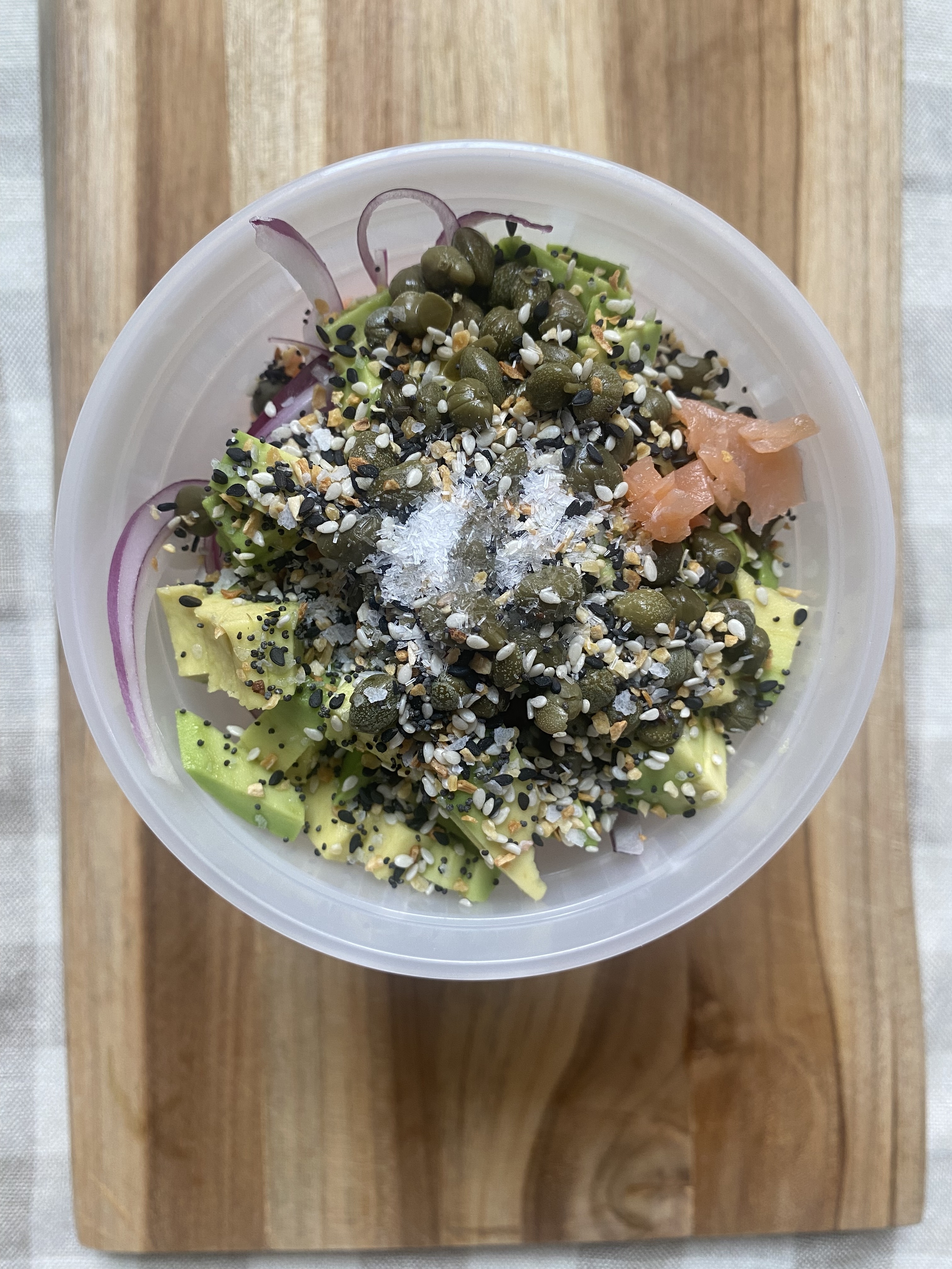A plastic container contains avocado, red onion, capers, sesame seeds, and smoked salmon