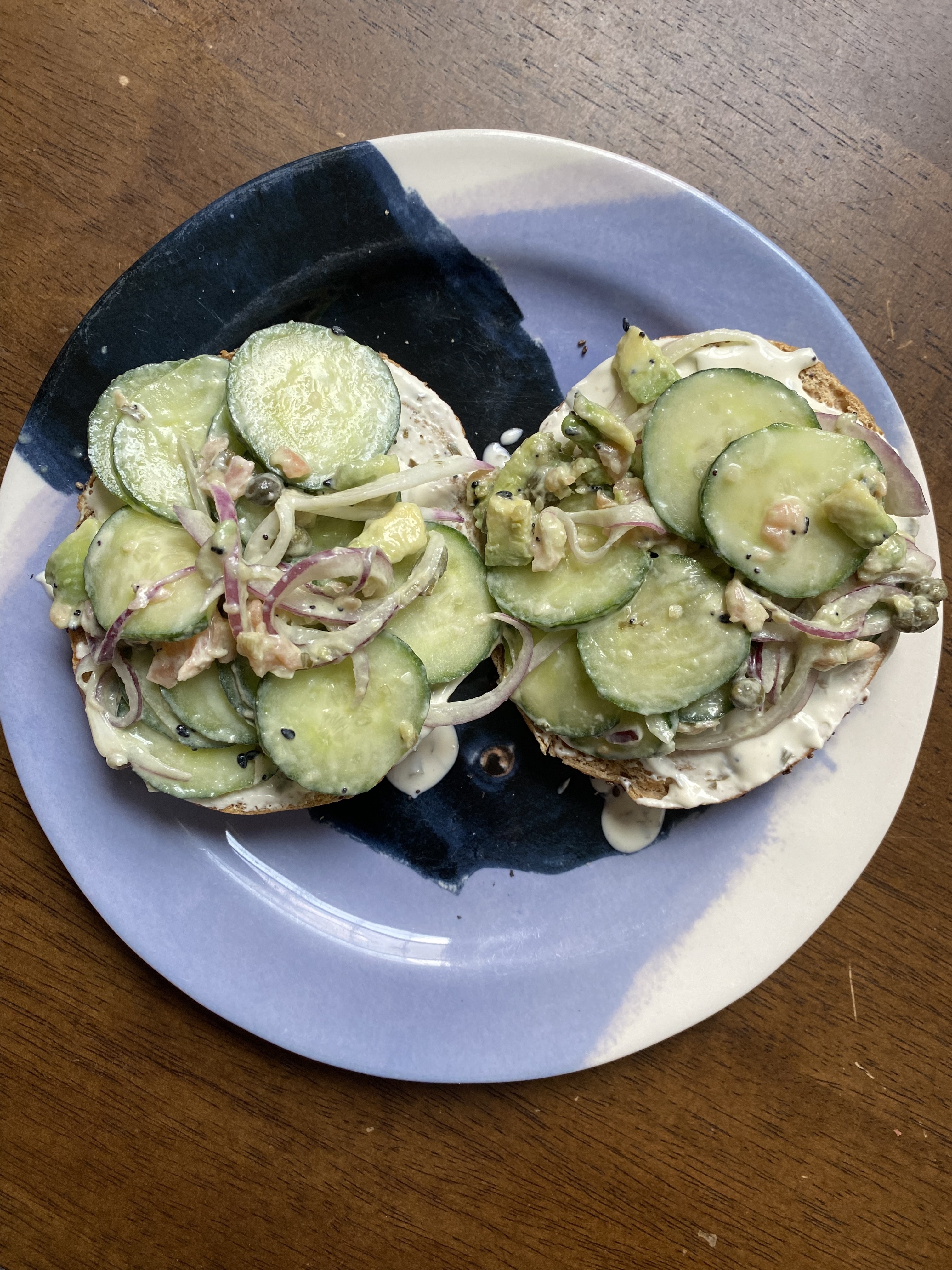 Two slices of bagel topped with cucumber slices, salmon, red onions, and cream cheese on a plate