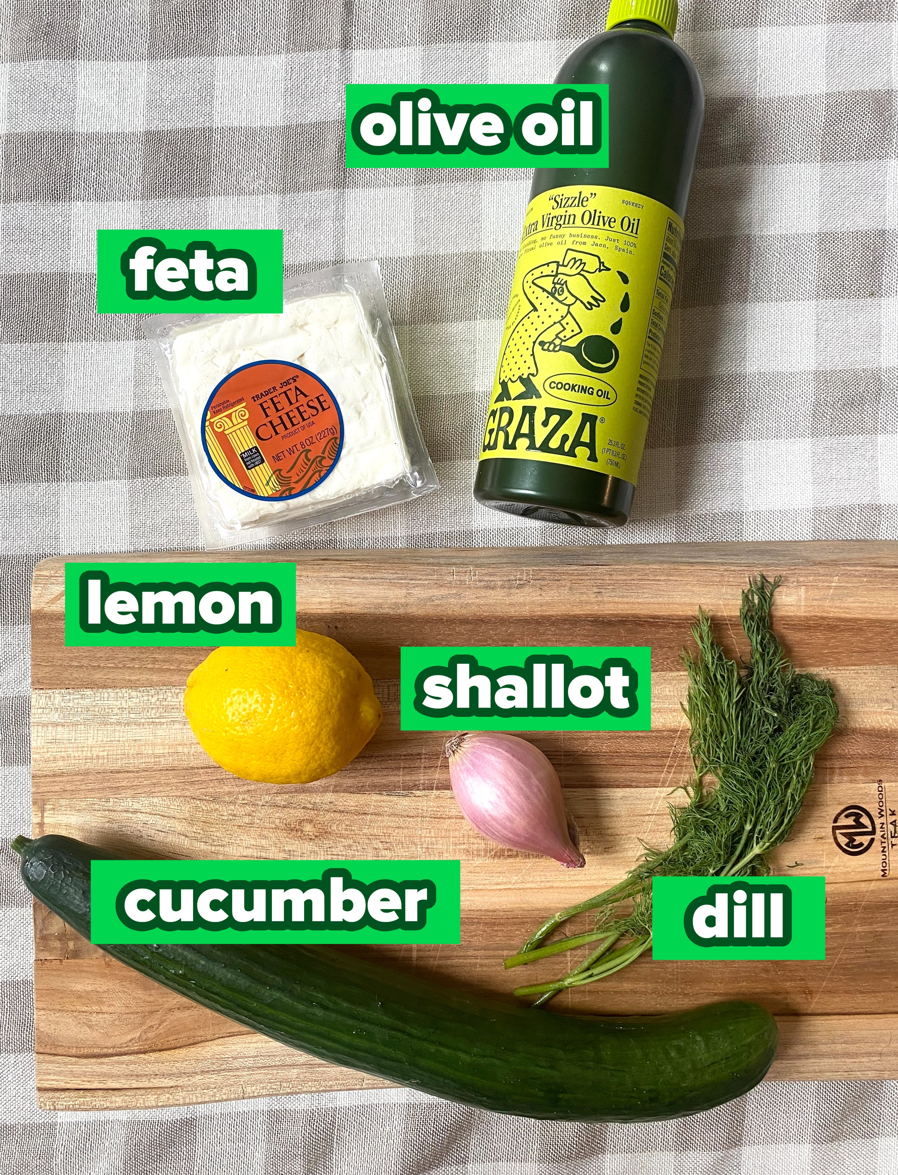 A cucumber, a lemon, a shallot, dill, a block of feta cheese, and a bottle of extra virgin olive oil on a cutting board