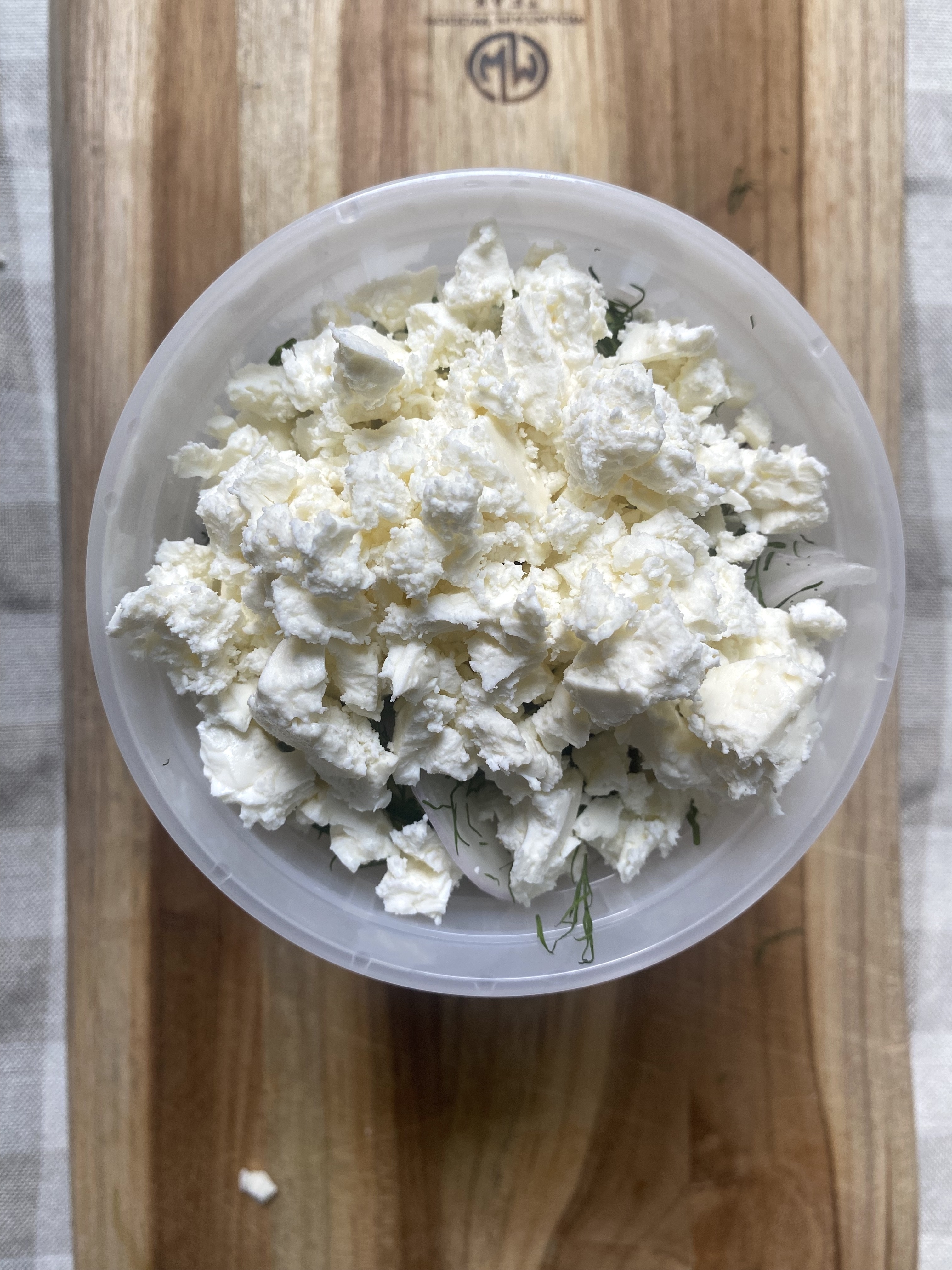 Plastic container filled with crumbled feta cheese, placed on a wooden cutting board