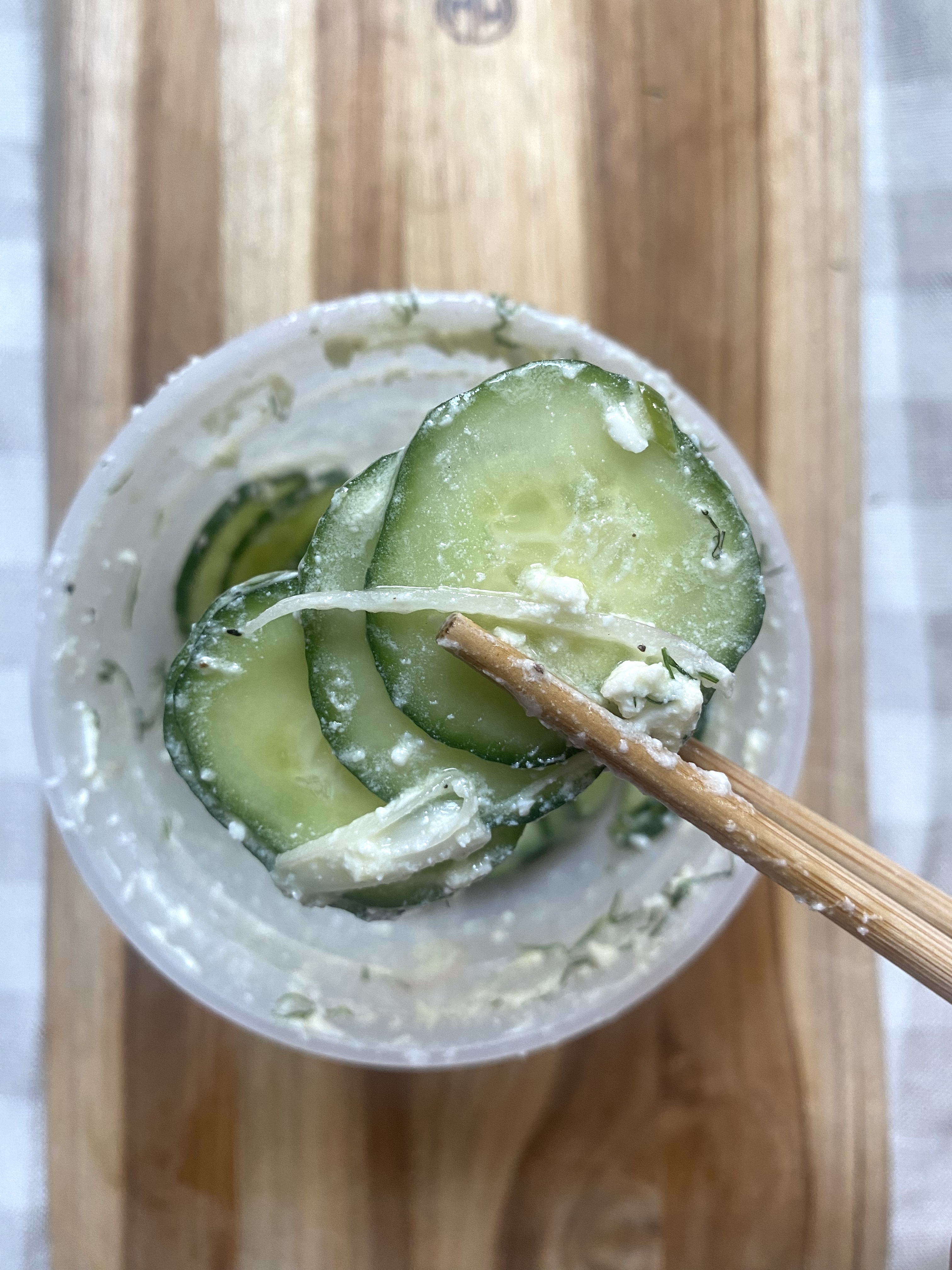 Slices of cucumber in a creamy, likely dill-flavored dressing, held by chopsticks over a container, placed on a wooden surface