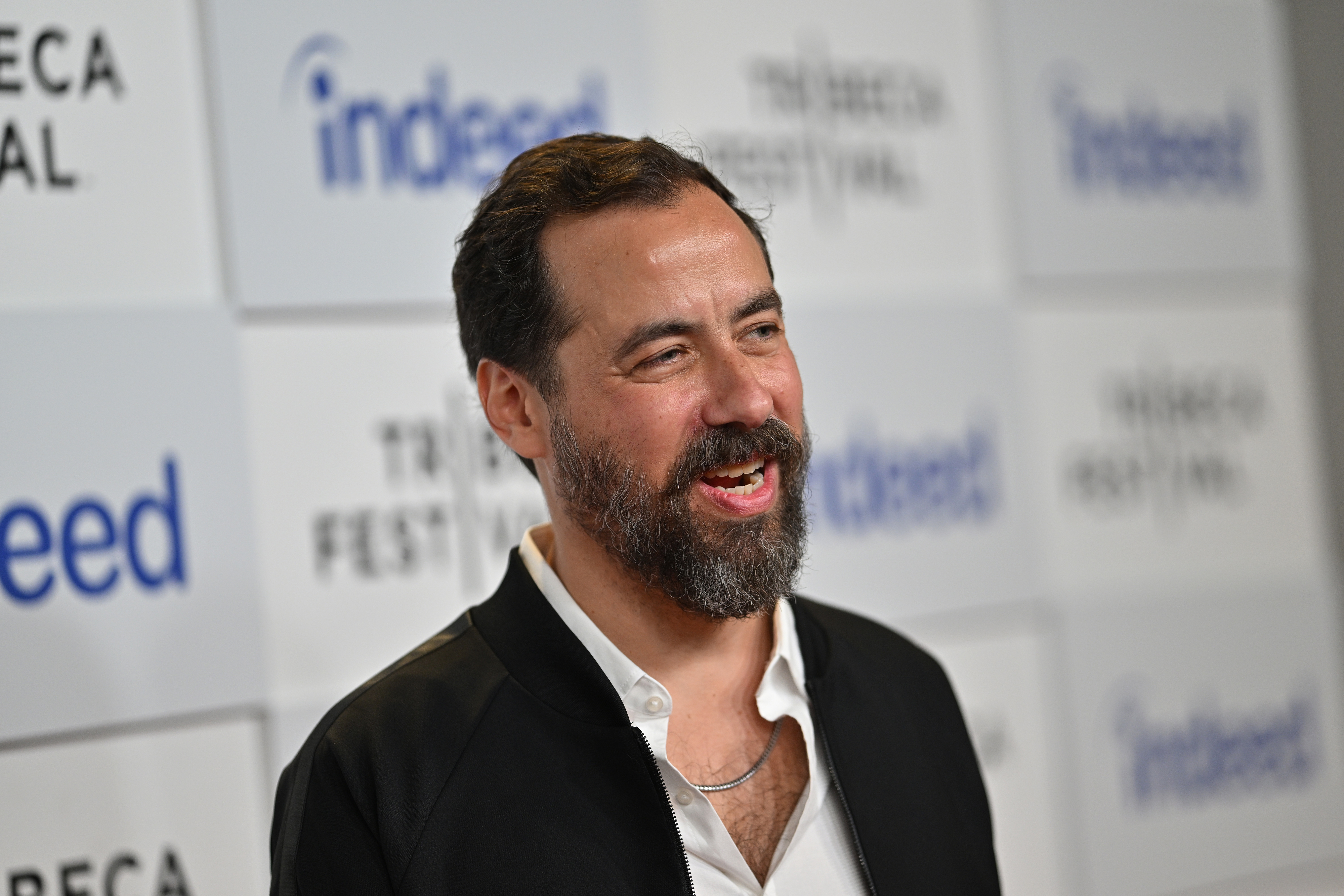 Eric Weinrib wearing a white shirt under a black jacket, stands smiling in front of a Tribeca Festival and Indeed event backdrop