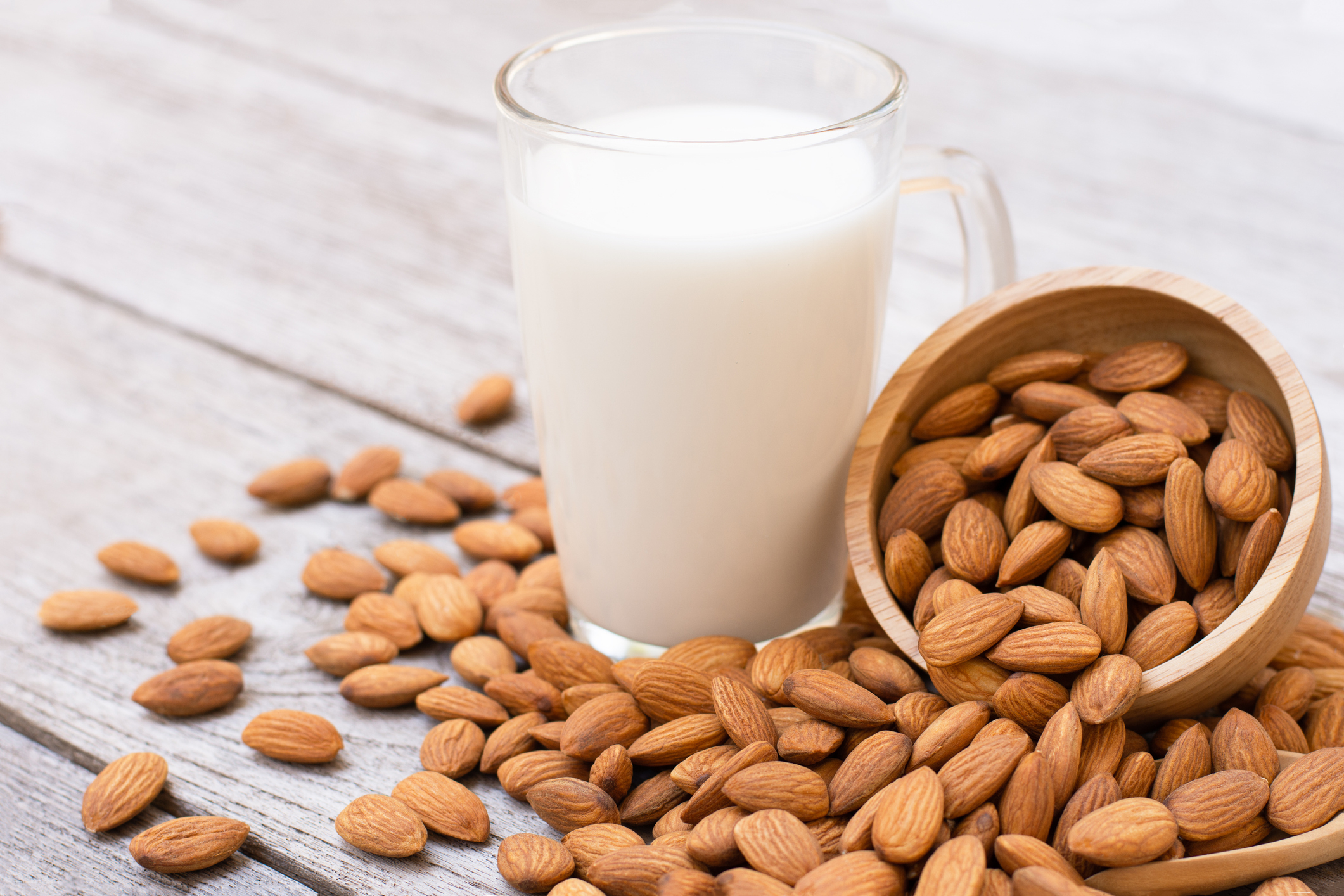 A glass of almond milk next to a wooden bowl tipped over with almonds spilling out onto a wooden surface