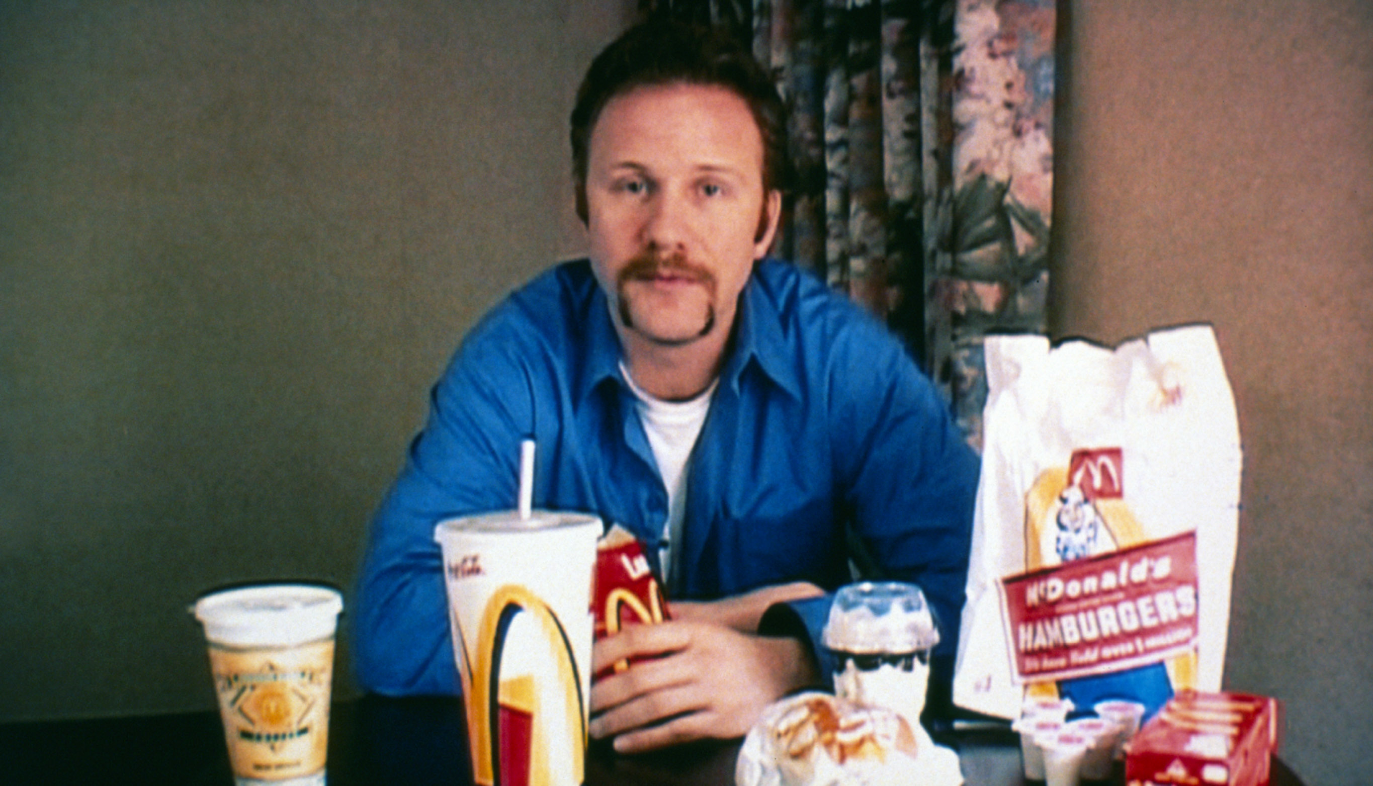 Morgan Spurlock sits at a table with various McDonald's items including drinks, burgers, and fries