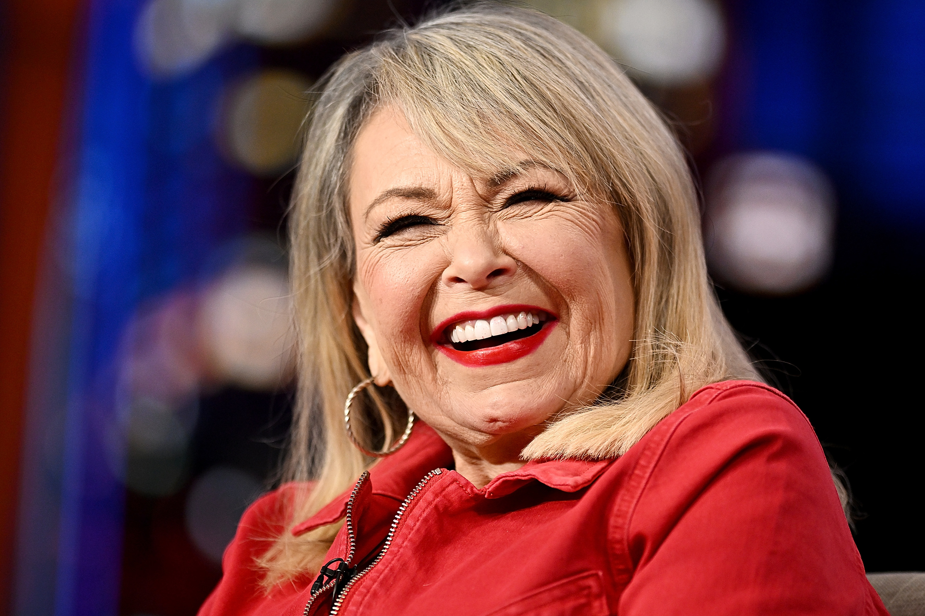 Roseanne Barr smiles broadly, wearing hoop earrings and a casual red jacket