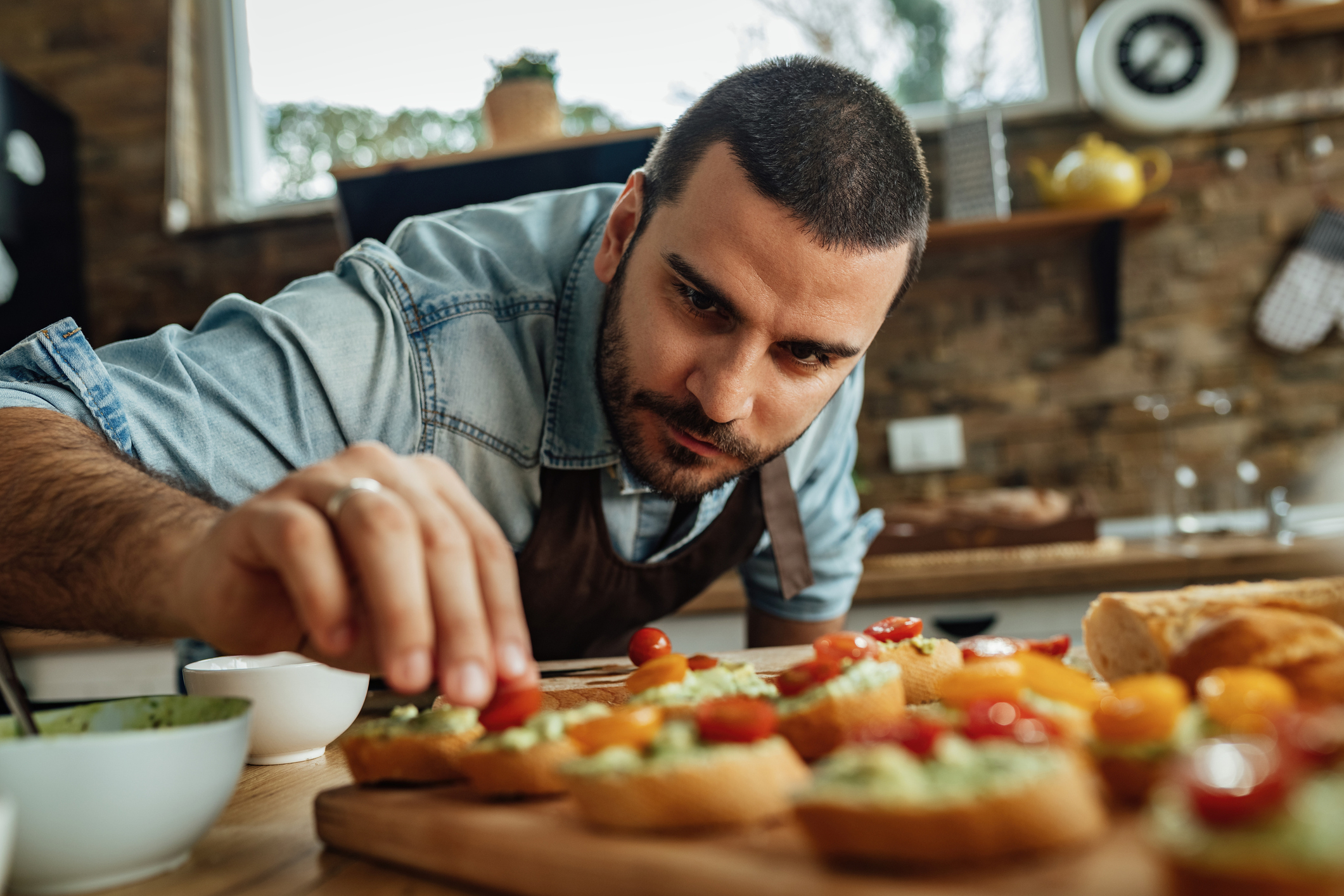A man in a casual shirt and apron carefully garnishes bruschetta in a modern kitchen, showcasing culinary skills and attention to detail