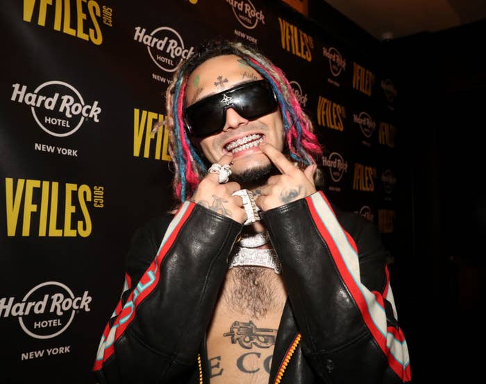 Lil Pump in a black jacket, wearing sunglasses, adorned with colorful dreadlocks, and posing with his fingers on his grin at a VFILES and Hard Rock Hotel event