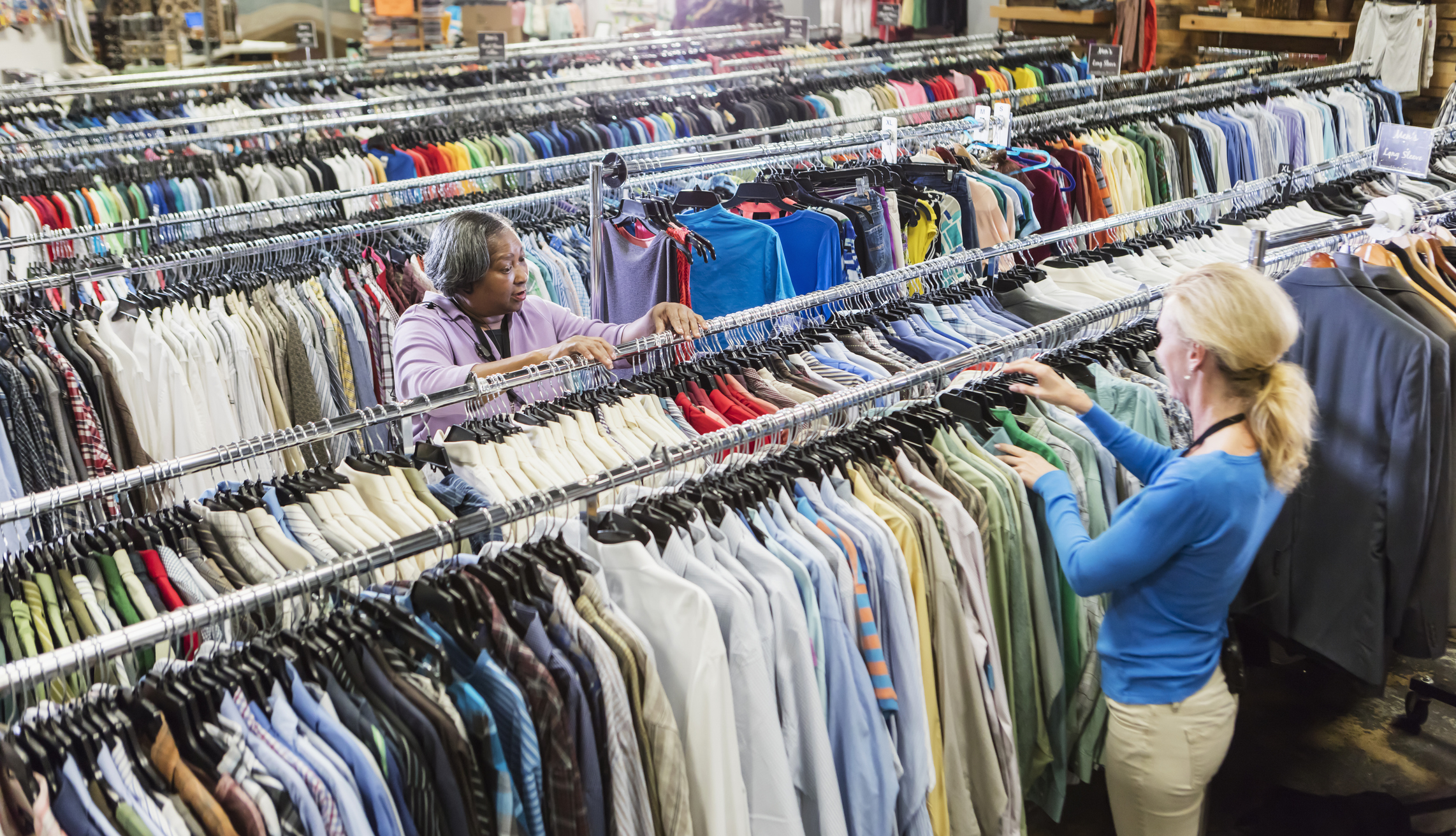 Two people sorting clothes on racks in what appears to be a thrift store or warehouse. The image relates to work and money aspects of clothing retail