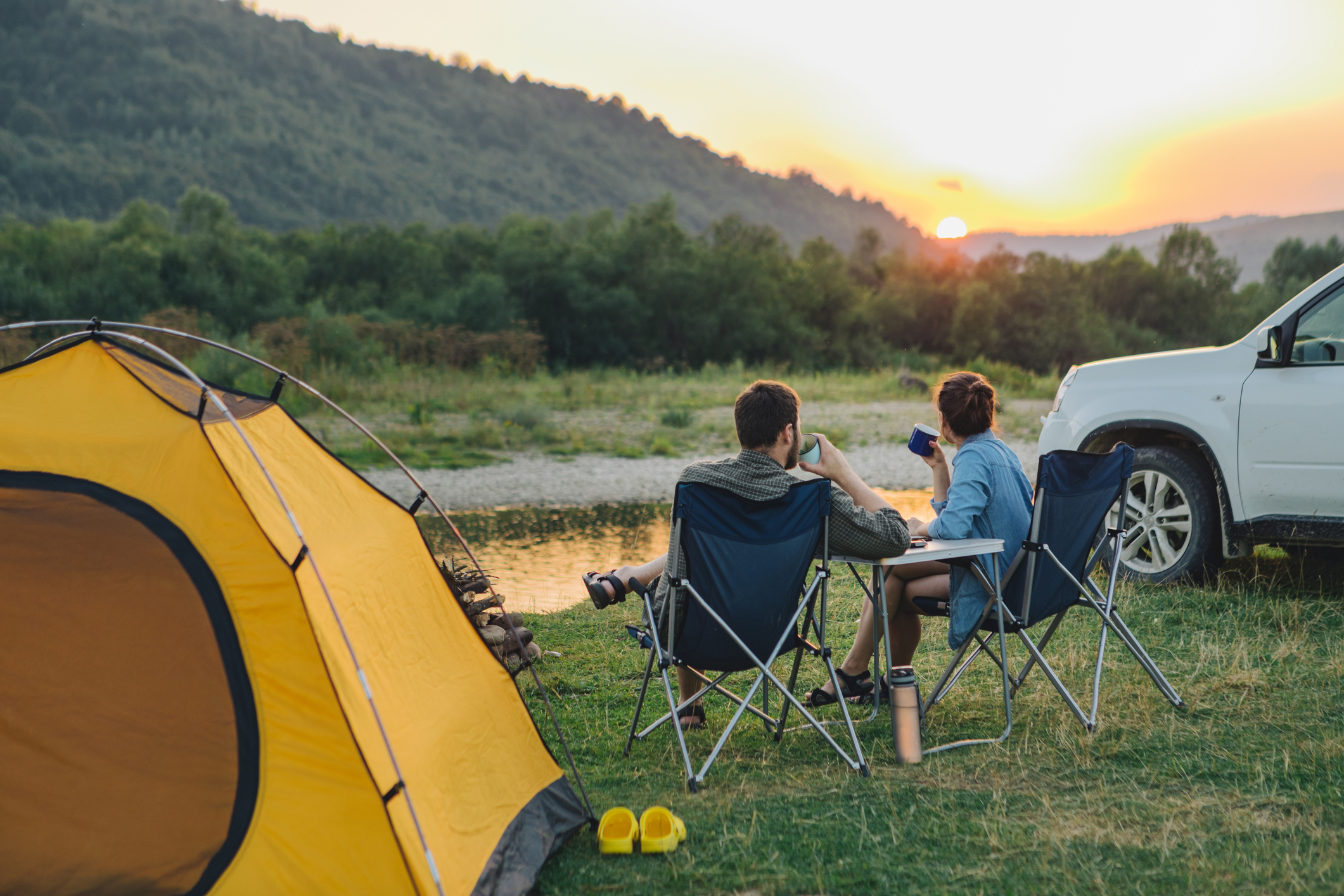 A man and woman sit in camping chairs near a tent and car, facing a sunset over a river and wooded hills