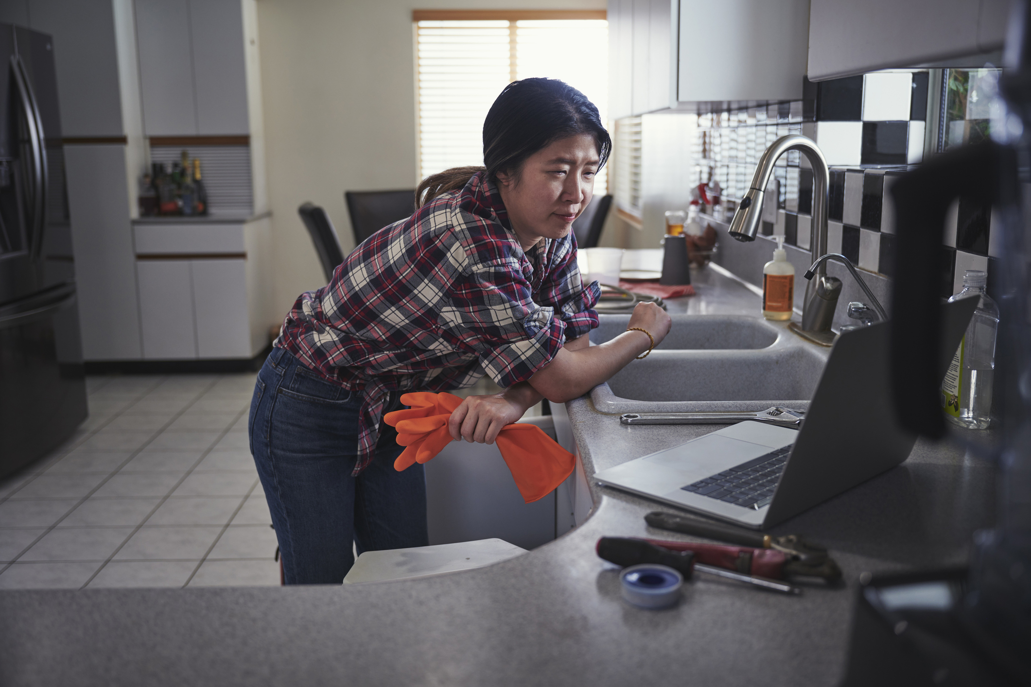 A person in a plaid shirt leans against a kitchen counter, watching something on a laptop. Cleaning gloves and a wrench are on the counter