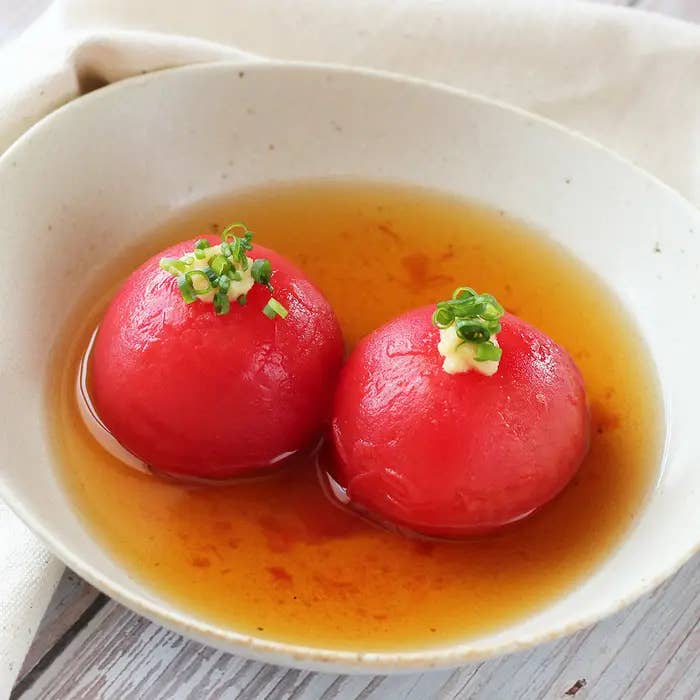 白い皿に盛り付けられた2つのトマトがだし汁に浸り、上にネギとおろししょうがが乗せられた料理。