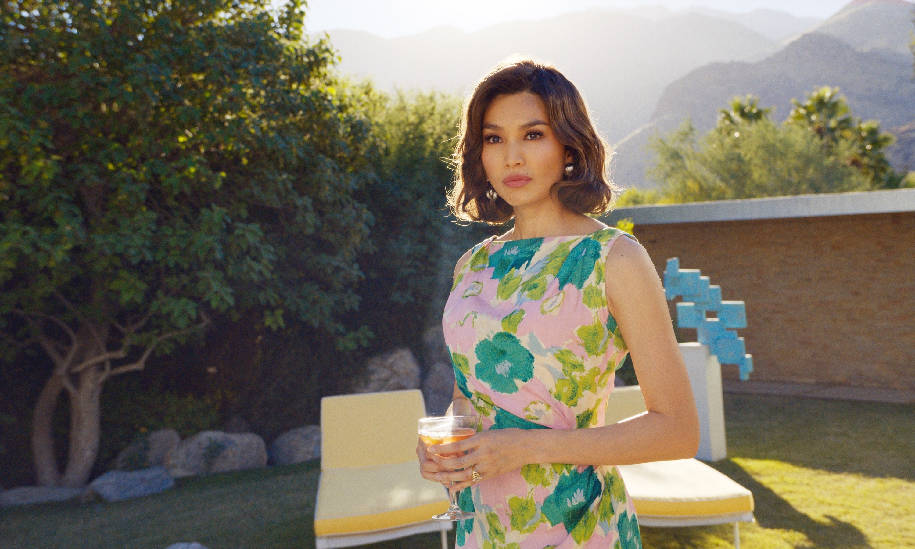 Gemma Chan in a floral print dress holds a beverage, standing outdoors with scenic mountains and garden backdrop in "Don't Worry Darling"