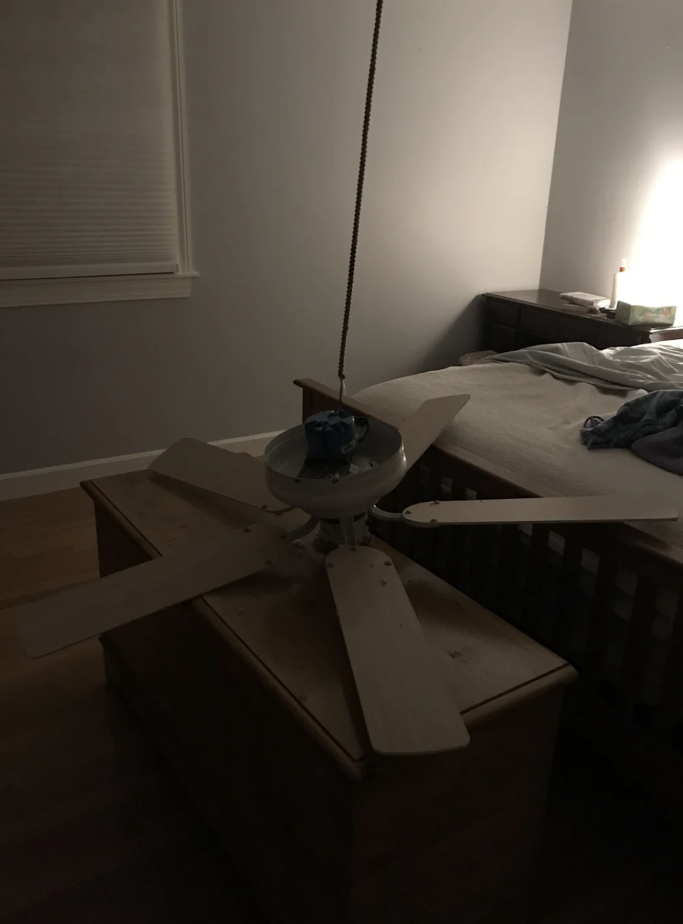 Bedroom with a ceiling fan detached and resting on a wooden chest at the foot of a bed. Dim lighting from a bedside lamp
