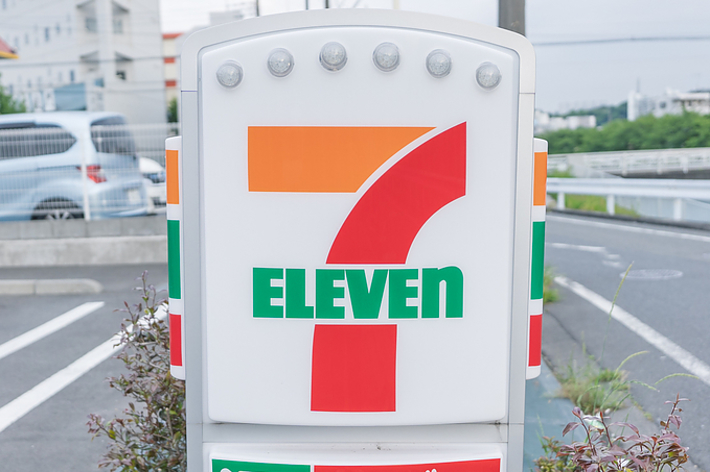 セブンイレブンの看板。アルコール、たばこ、銀行ATMのサービスを提供。背景に駐車場と建物がある。