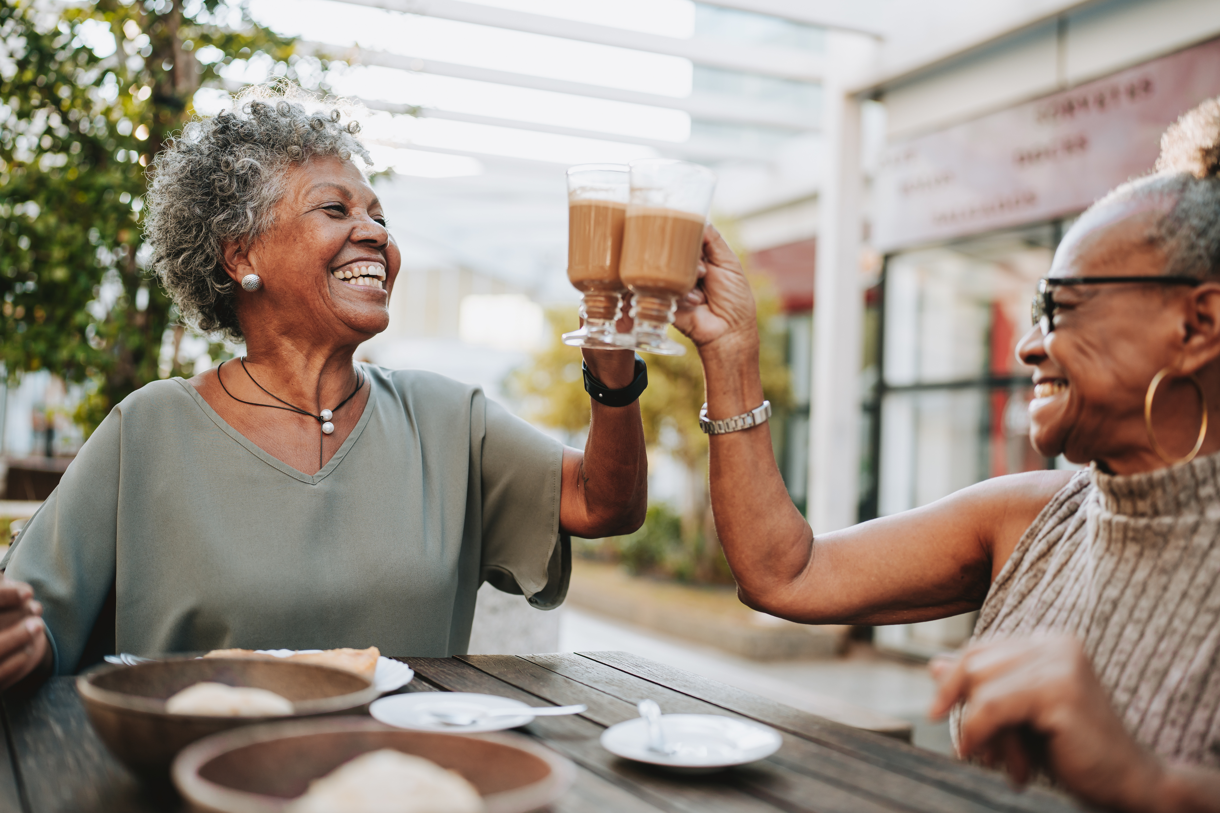 Two women smile and clink glasses at an outdoor café table, enjoying drinks and sharing a joyful moment