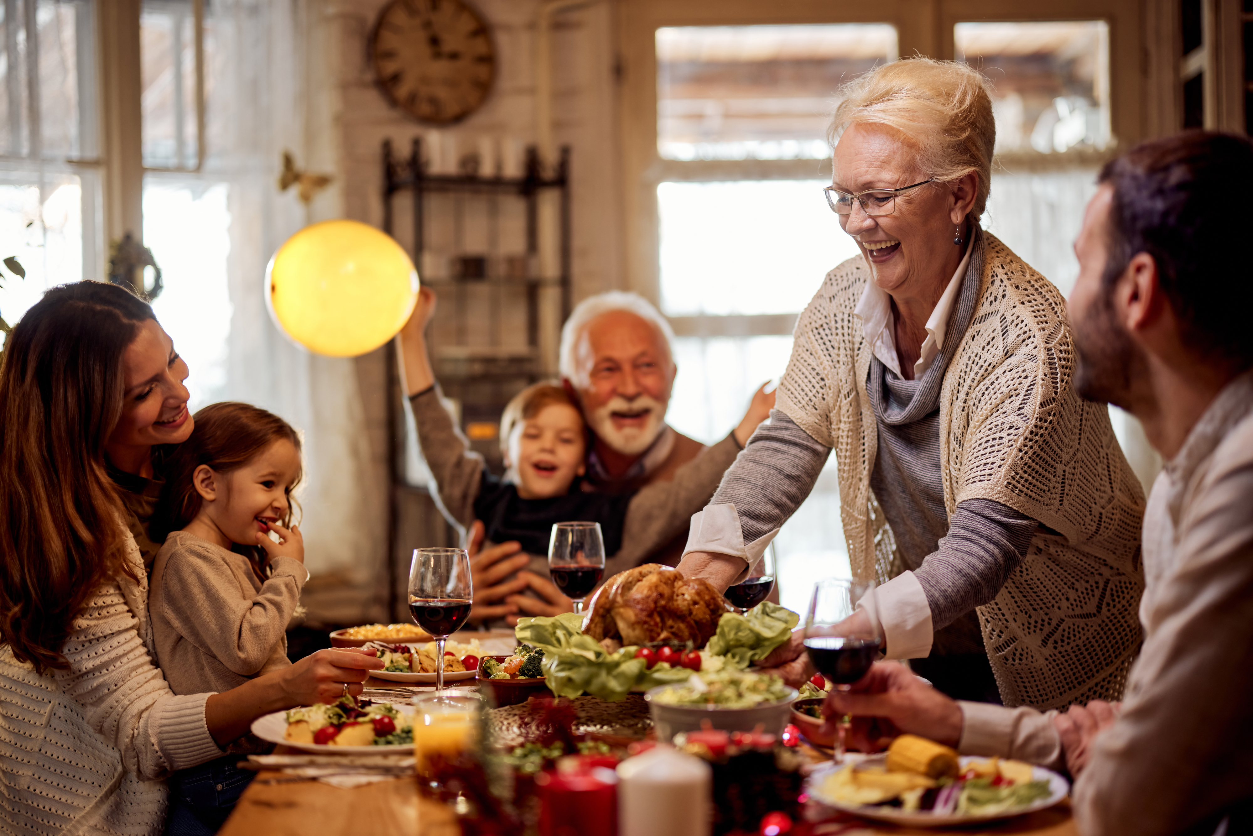 A smiling elderly woman serves a roast chicken to a joyful family of six seated around a festive dining table