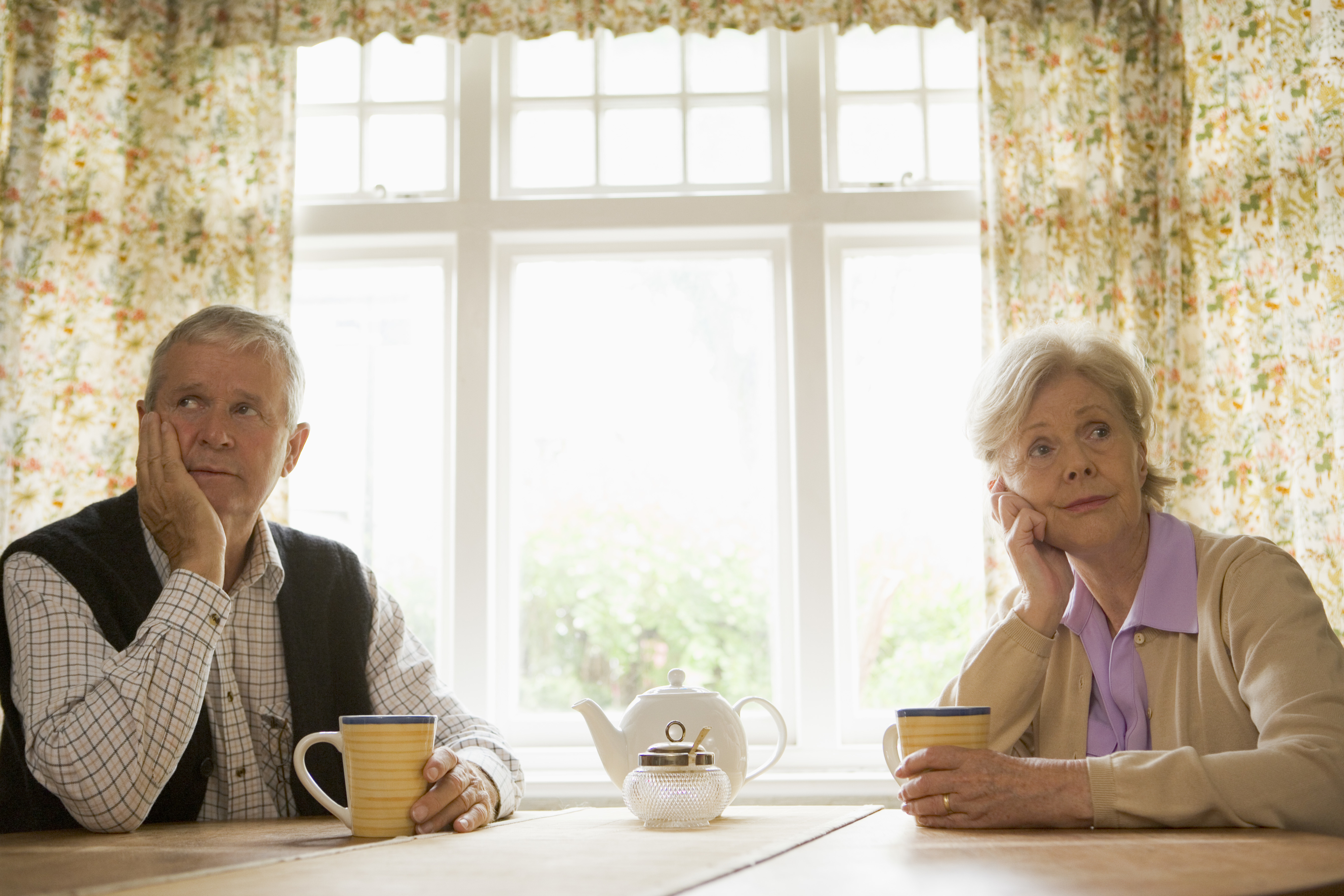 An elderly man and woman sit at a table holding cups of tea, looking contemplative. A teapot is on the table between them