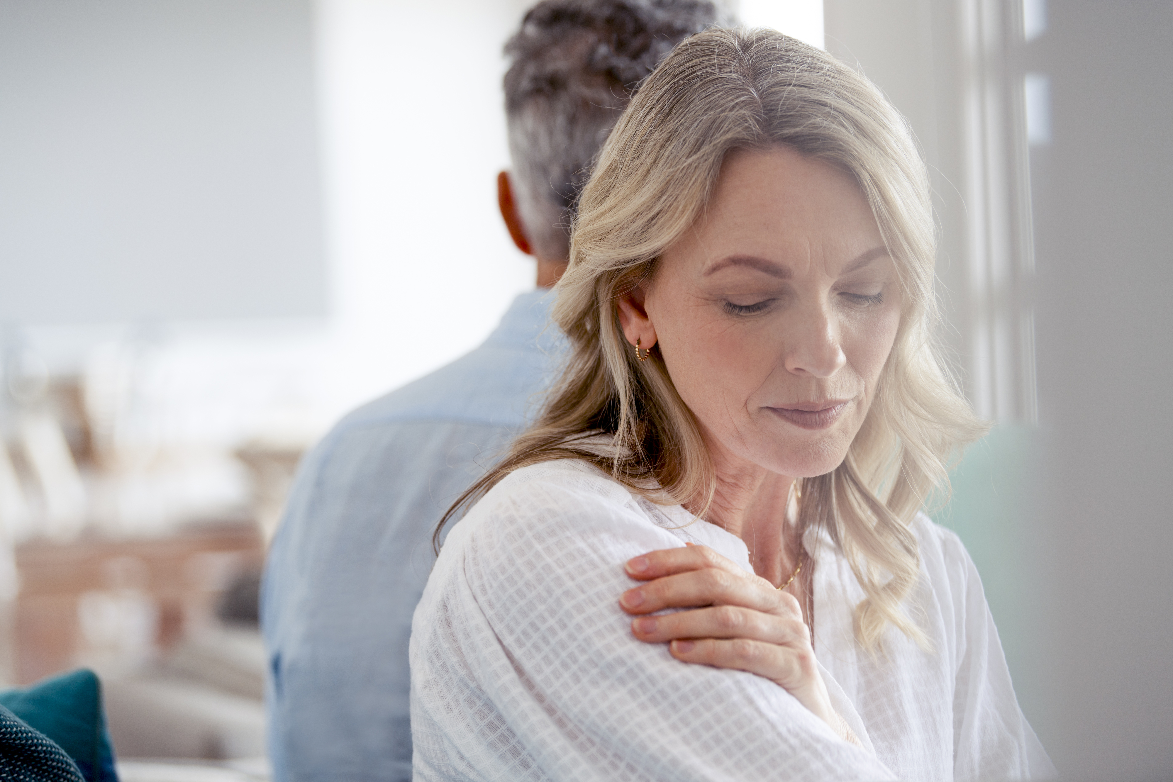 A woman and a man sit back-to-back, the woman looking down with a thoughtful expression and gently holding her own shoulder