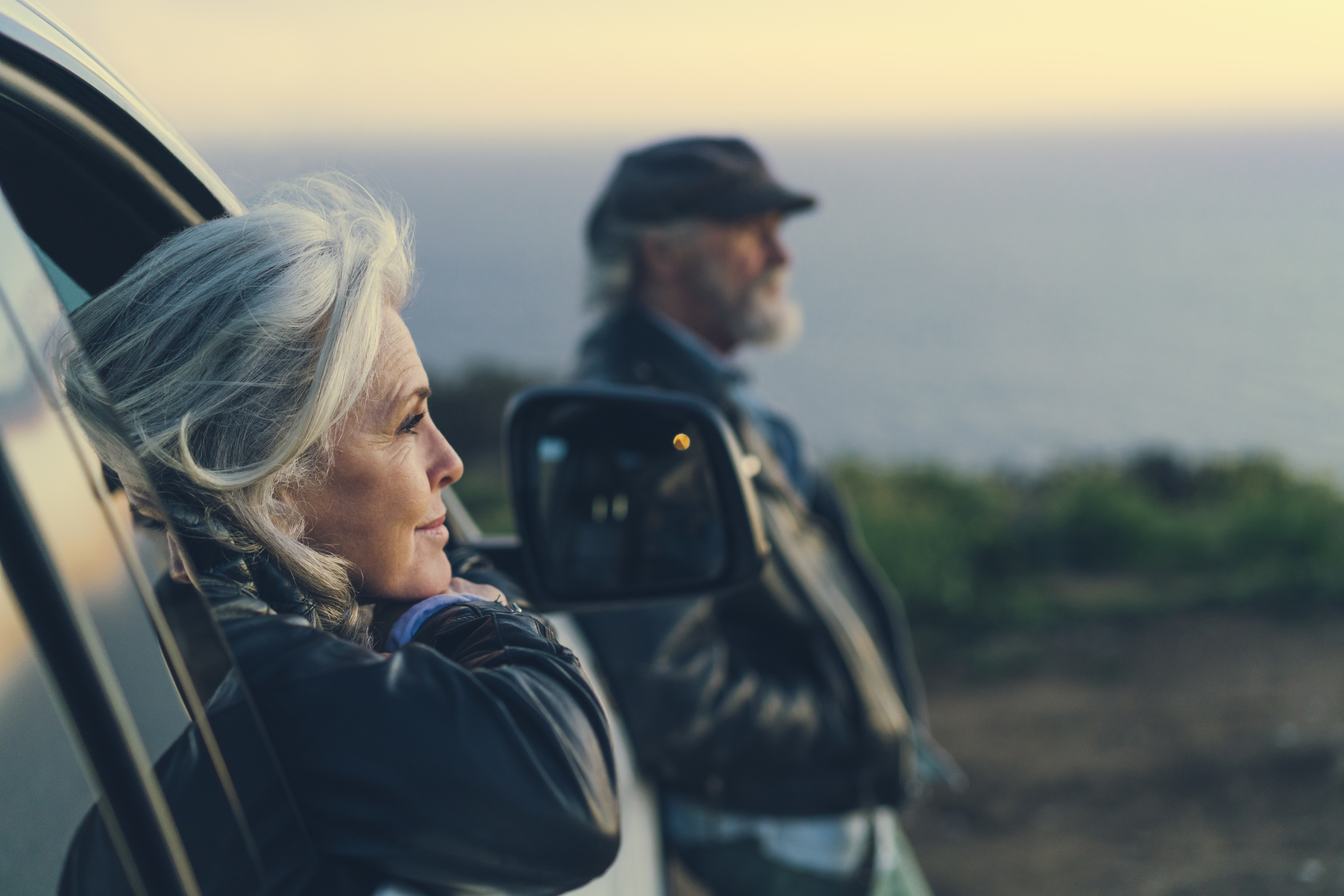 An older woman with gray hair leans against a car door looking out into the distance. An older man stands nearby also gazing into the distance