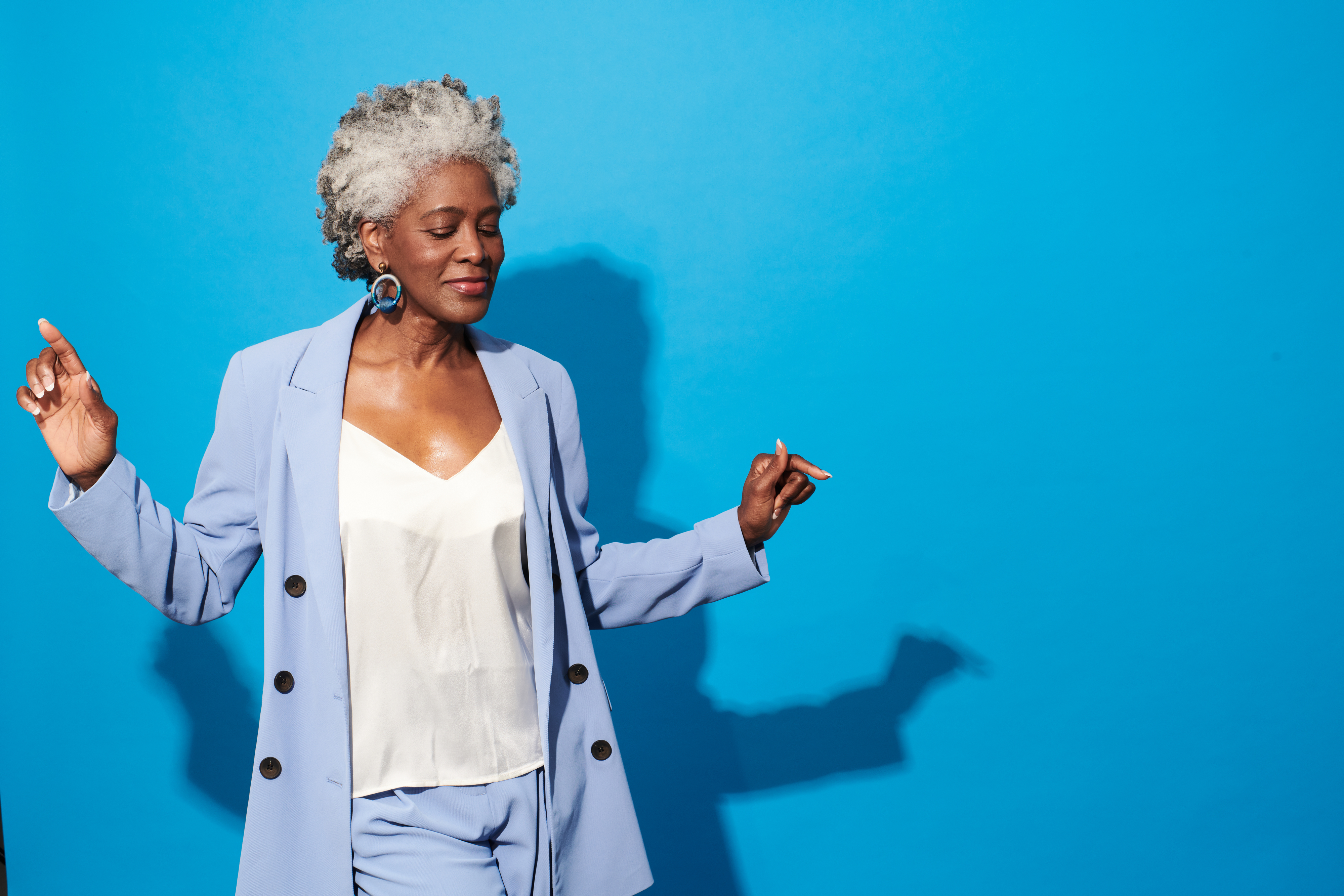 An older woman with curly gray hair dances joyfully. She is wearing a light-colored suit with a white blouse and large earrings