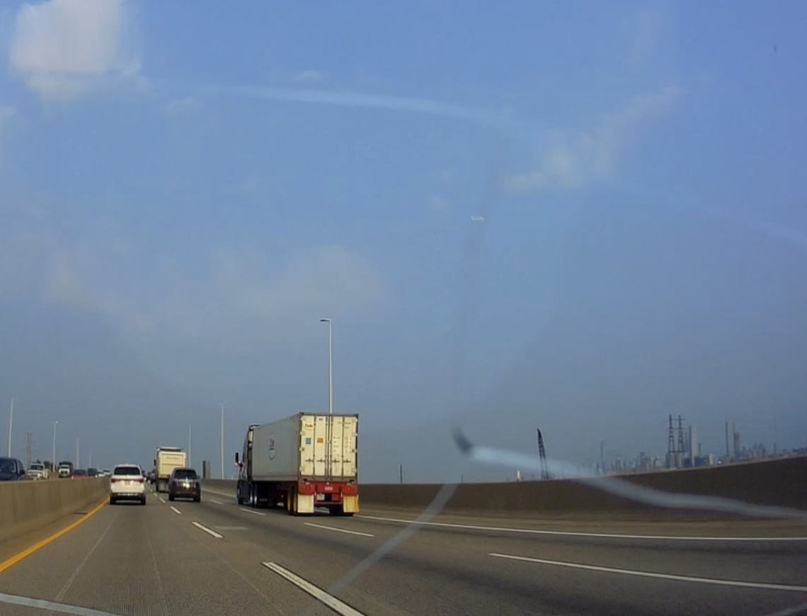 Vehicles driving on a multi-lane highway, including a white truck in the center lane and a white car on the left. An industrial area is seen in the background