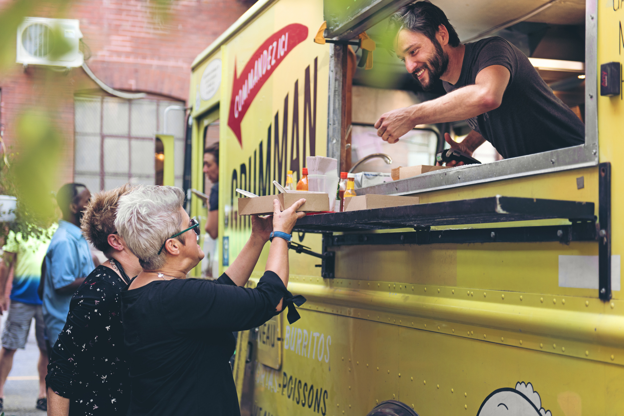 Keanu Reeves smiles while serving food from a truck to two women in a casual setting. The photo captures a friendly interaction at a food event