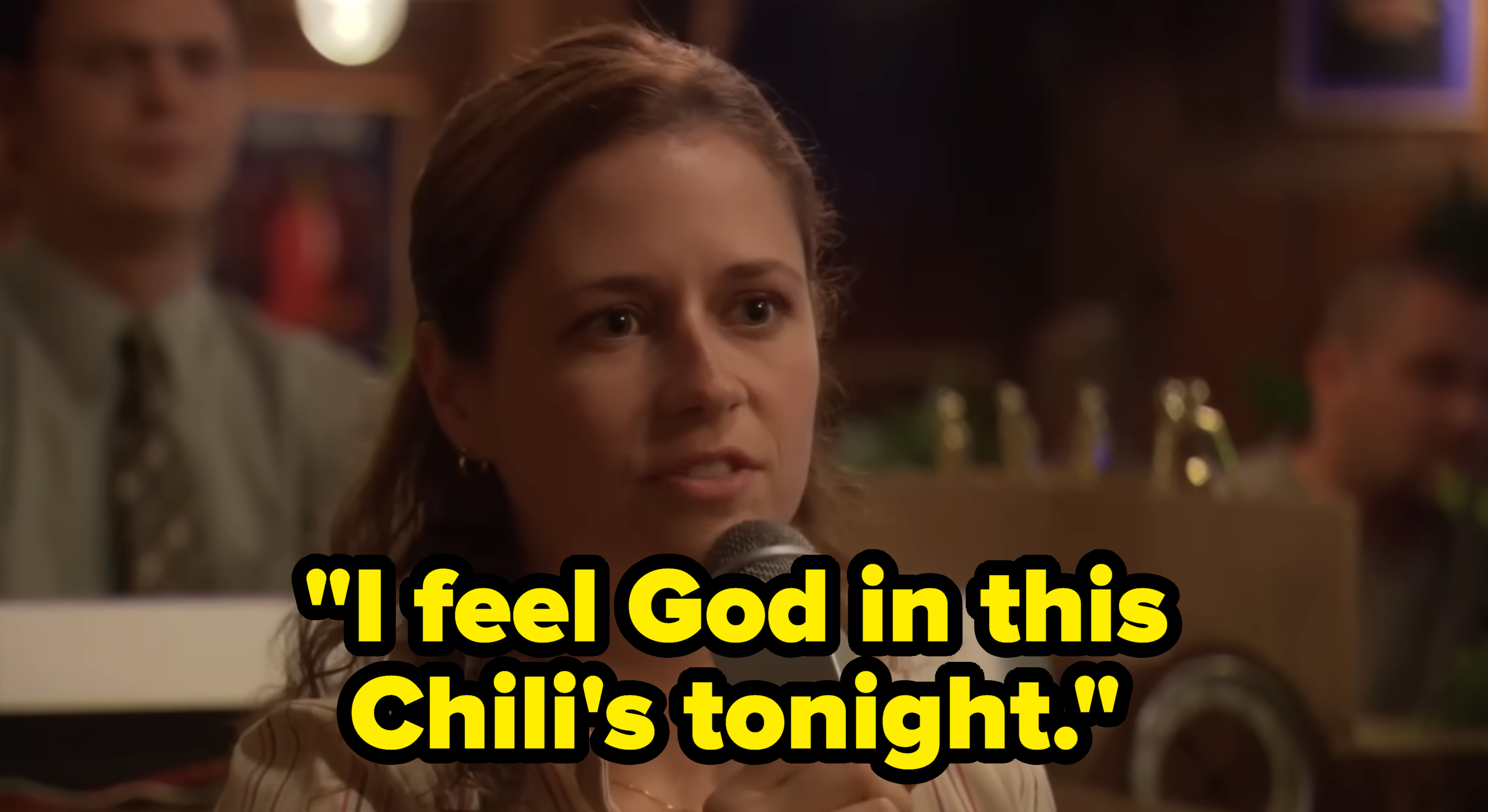 Jenna Fischer, holding a microphone, speaks with a pensive expression in a dimly lit setting. Text reads, "I feel God in this Chili's tonight."