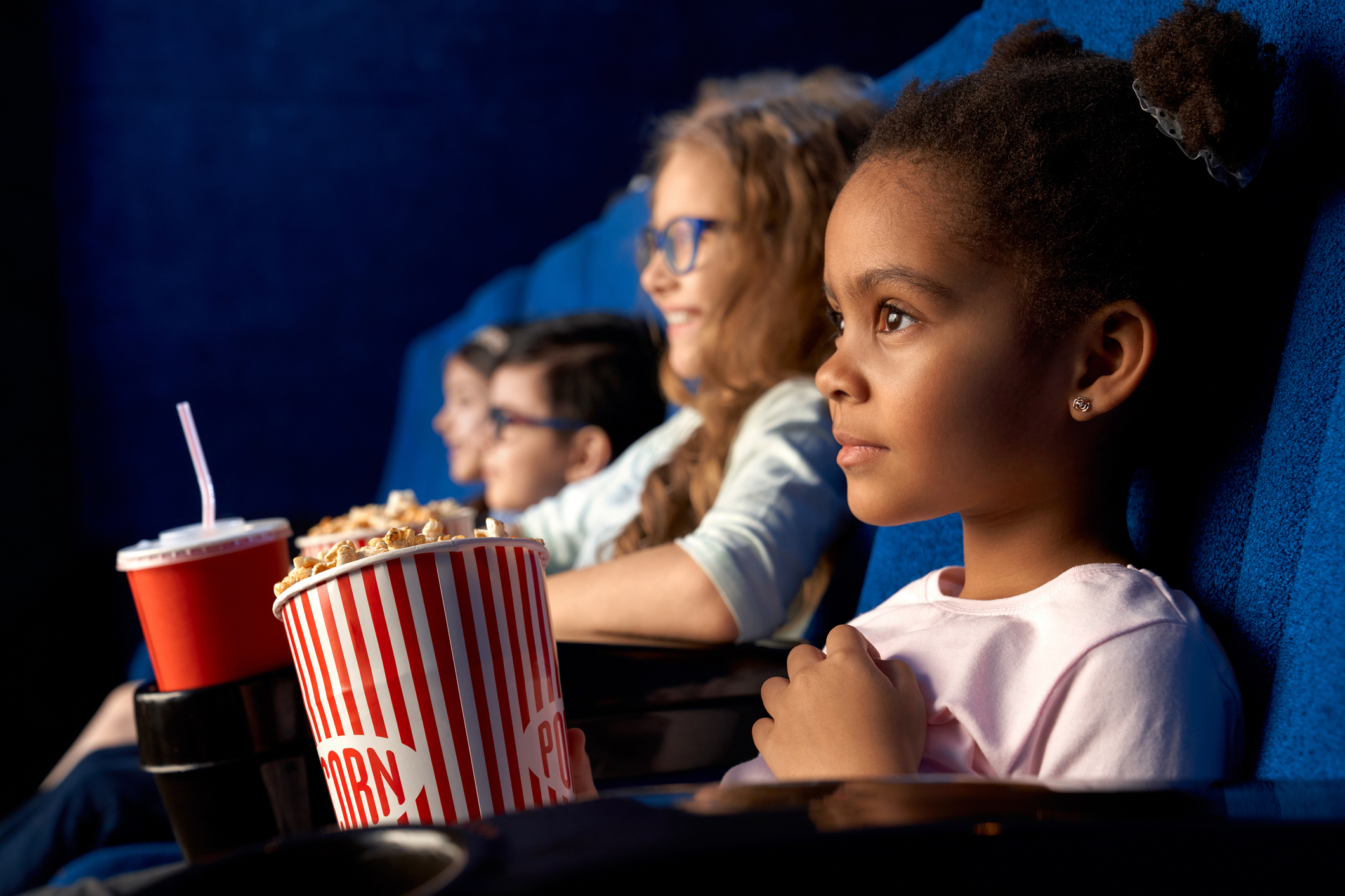 Four children watch a movie in a theater, holding popcorn and drinks. They are focused on the screen ahead
