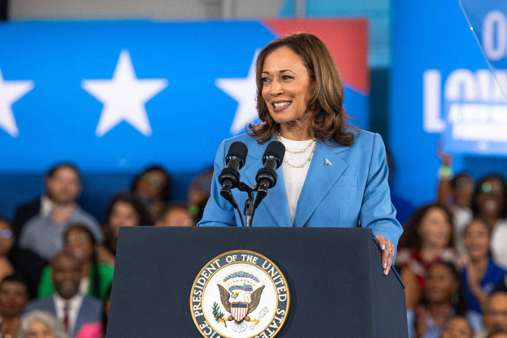 Kamala Harris stands at a podium with the U.S. Vice Presidential seal, addressing an audience. She wears a formal suit