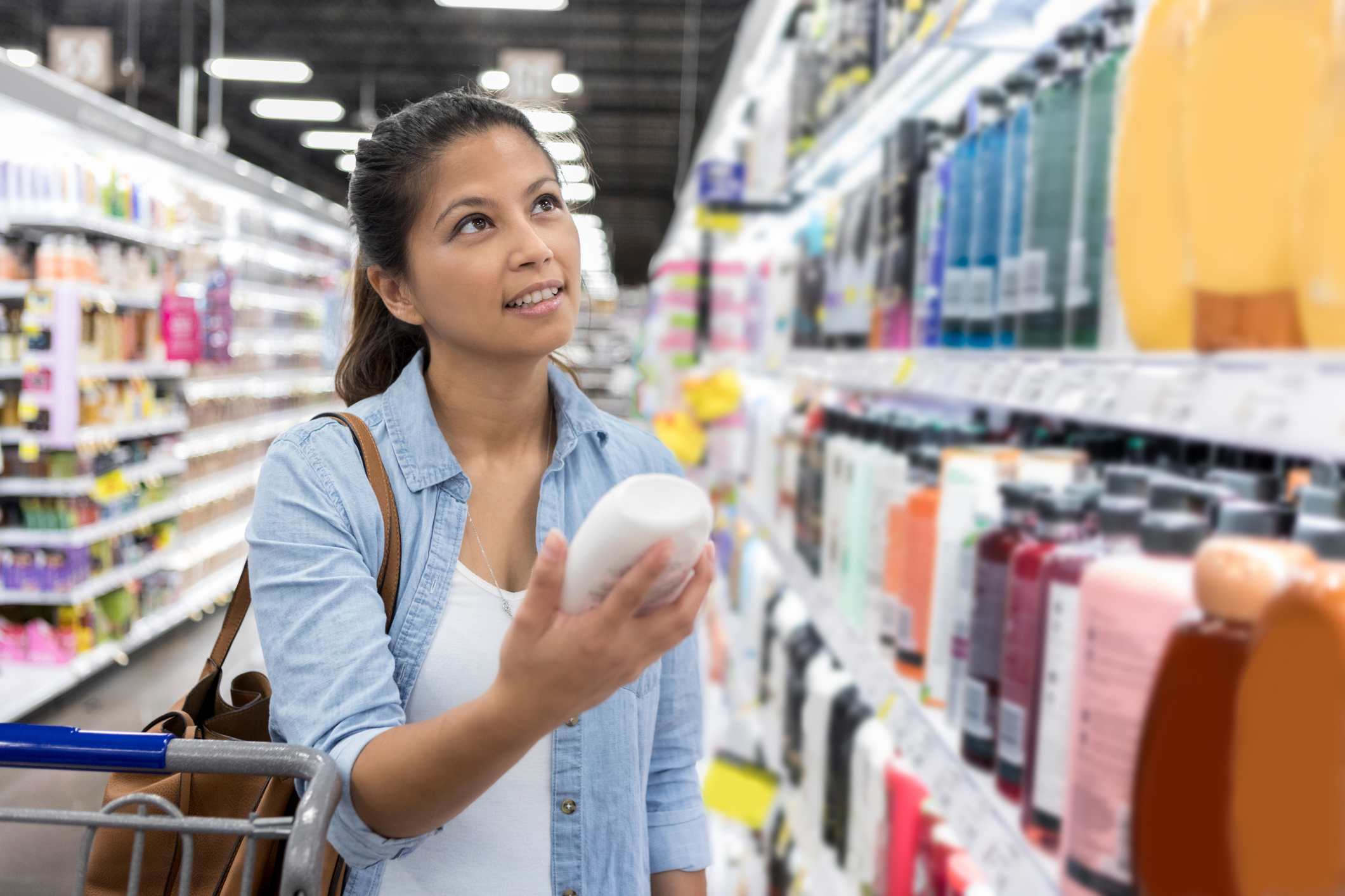 Woman shopping in an aisle with personal care products, holding a bottle and examining the label. Cart with groceries is beside her