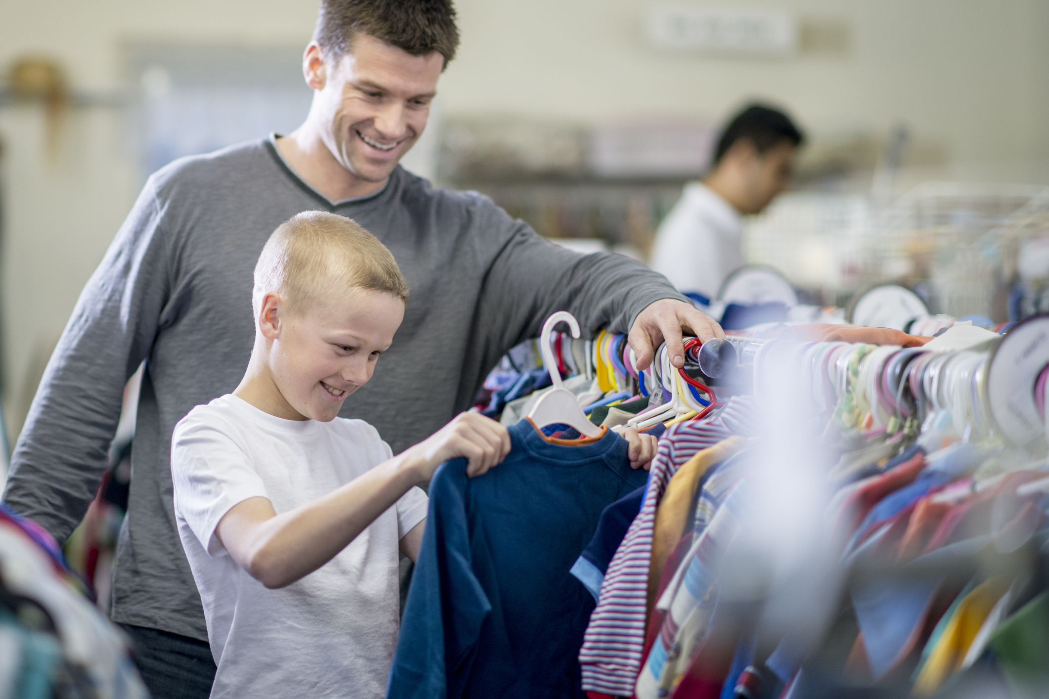A man and a boy are smiling while browsing through clothes on hangers in a thrift store. The boy is holding up a shirt, appearing excited about the selection