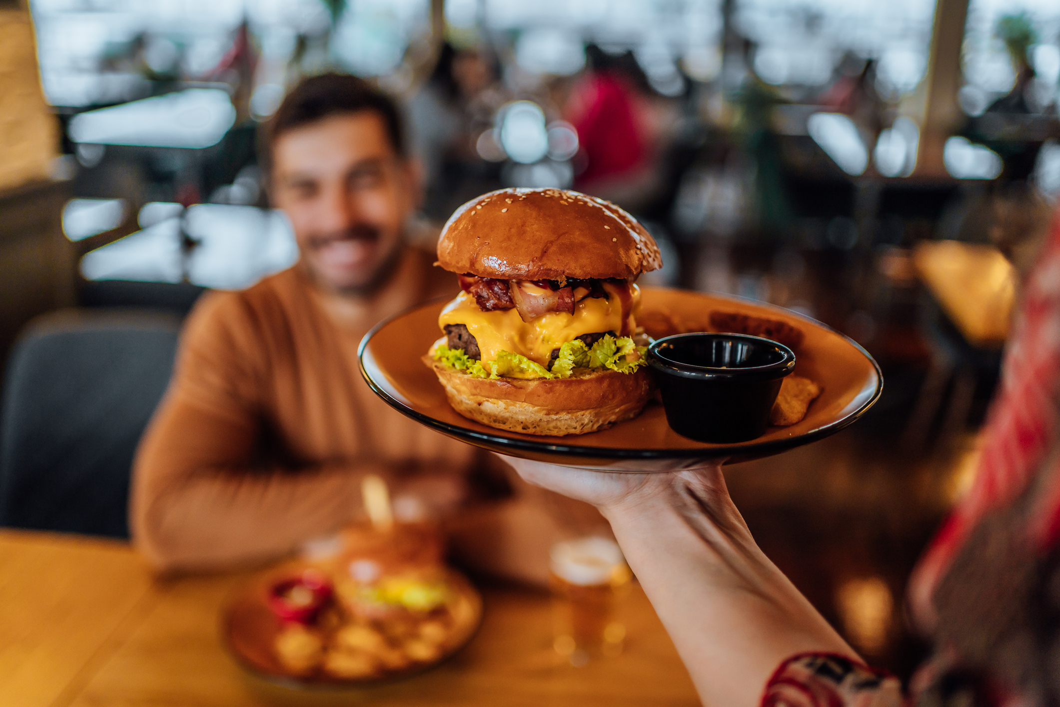 A server presents a burger with bacon and cheese to a smiling man seated at a restaurant table